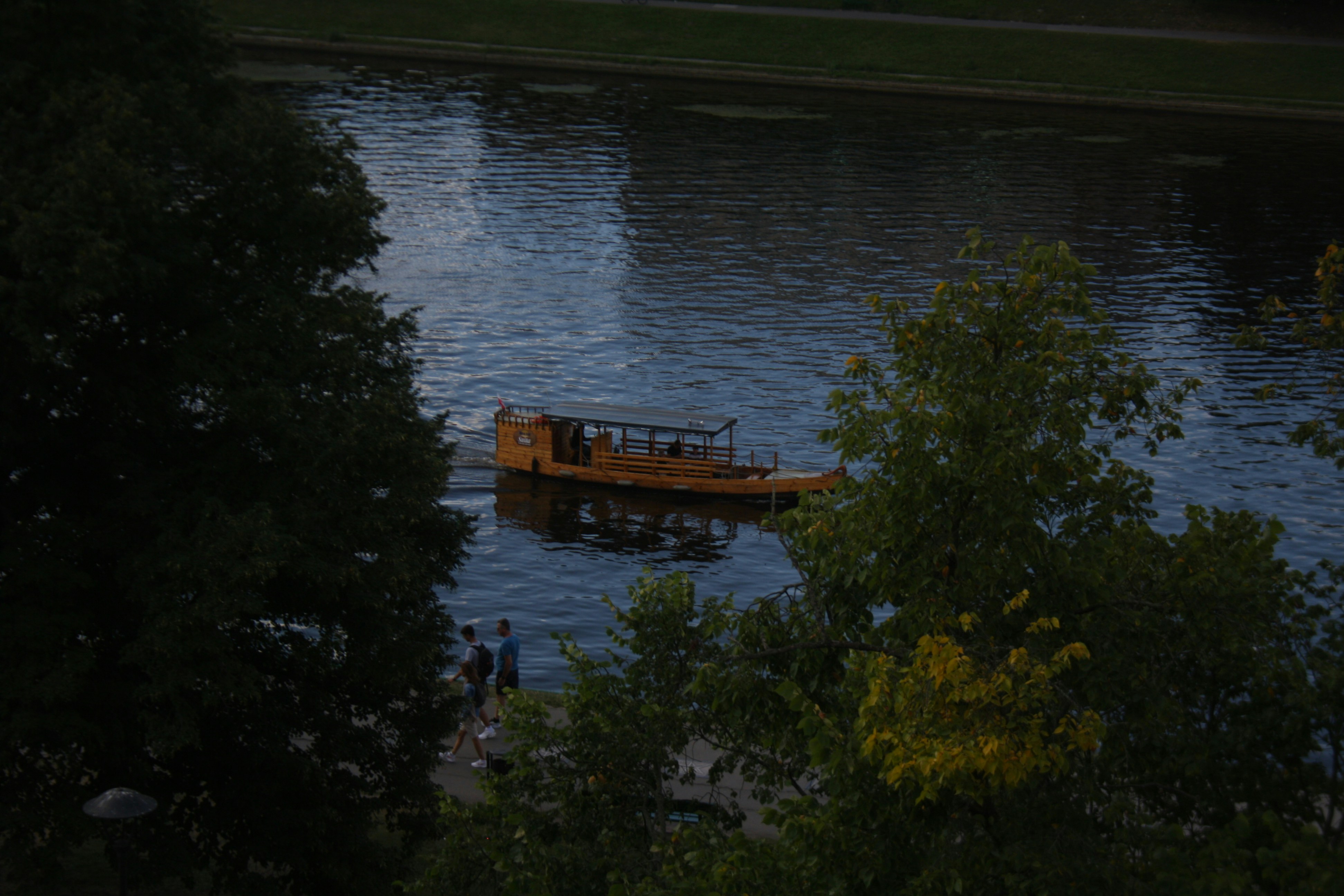 A wooden boat on a dark river near a tree-lined path with people