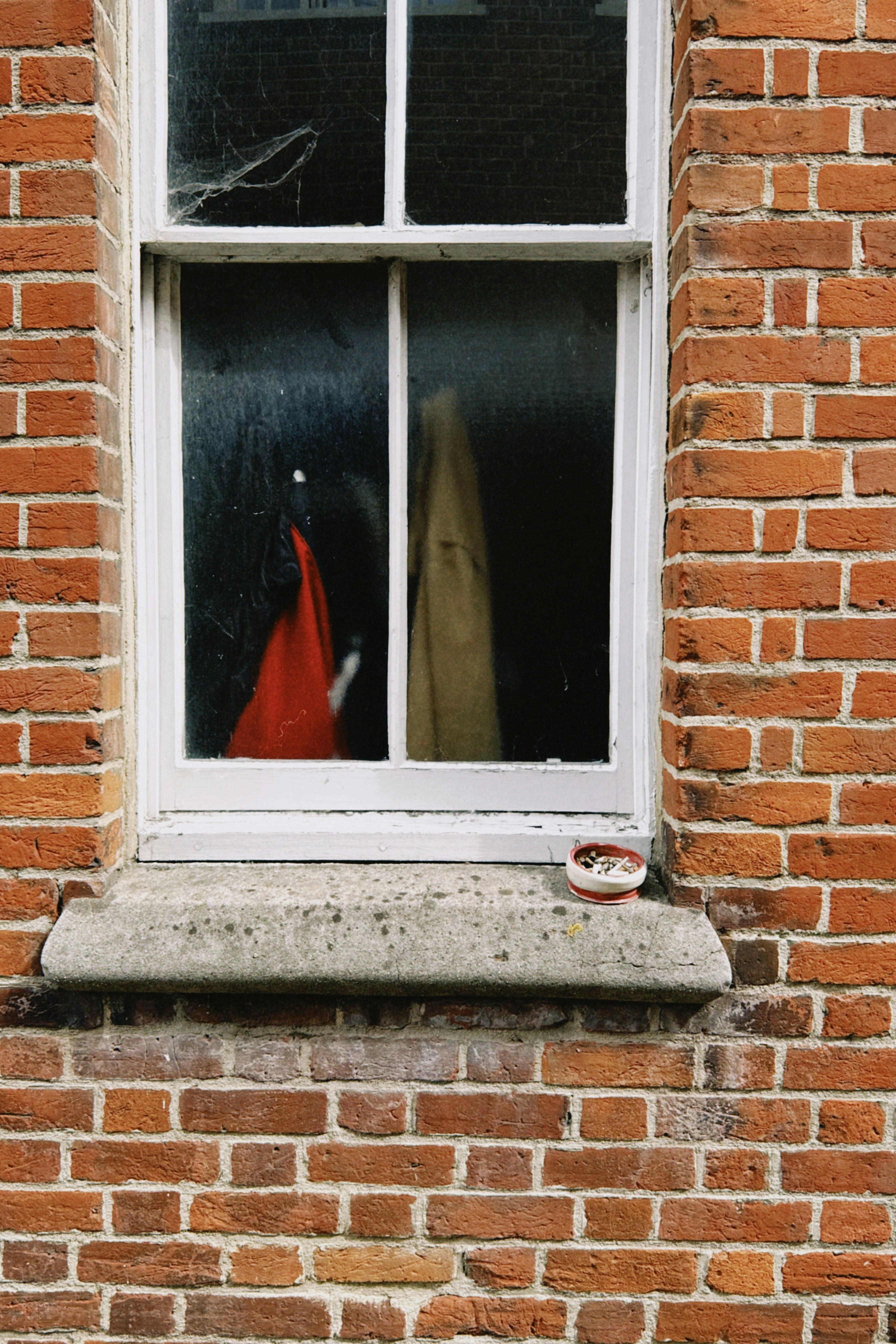 A red and a tan coat hanging inside a window with a brick wall