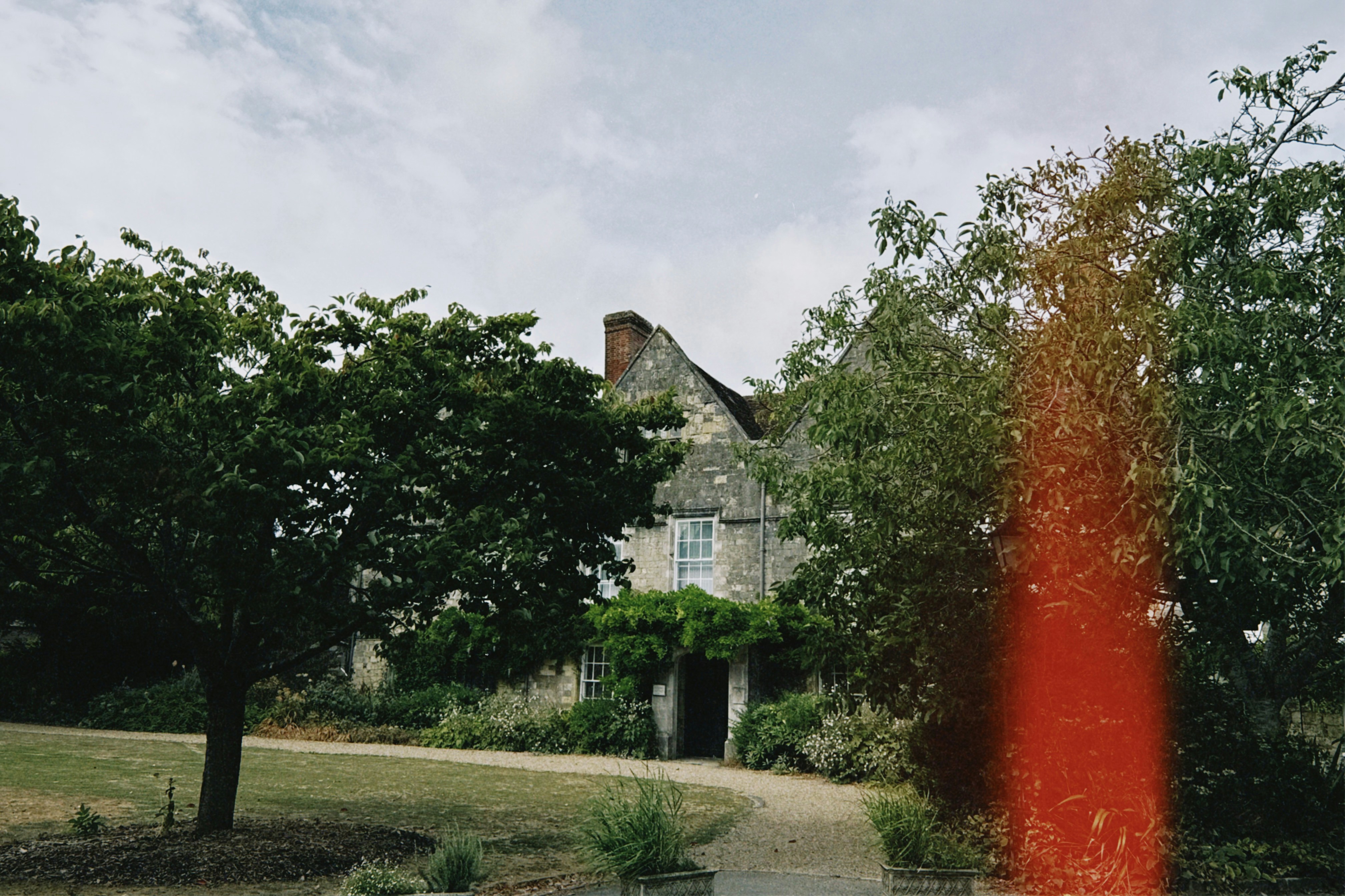 A stone manor house behind trees with a gravel path and light leak