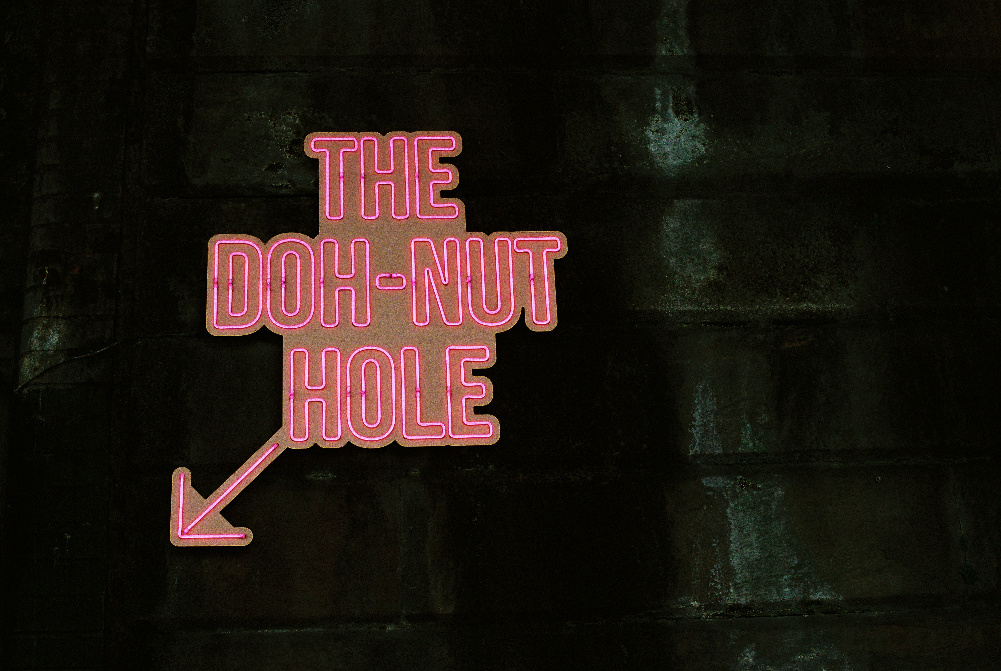 A pink neon sign reading THE DOH-NUT HOLE with an arrow on a dark wall