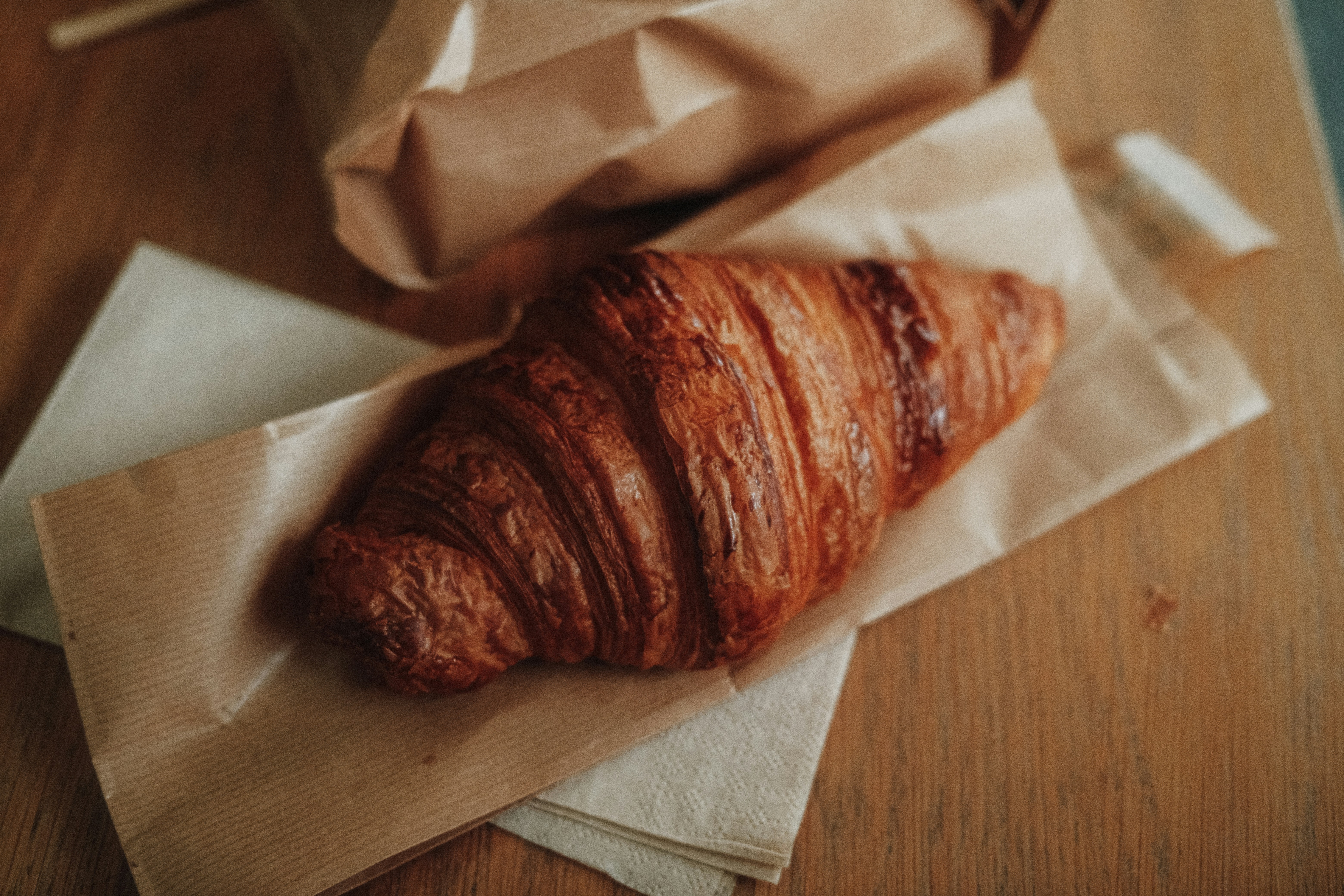 광명맘카페 - A golden brown croissant on a paper napkin and bag on a wooden table