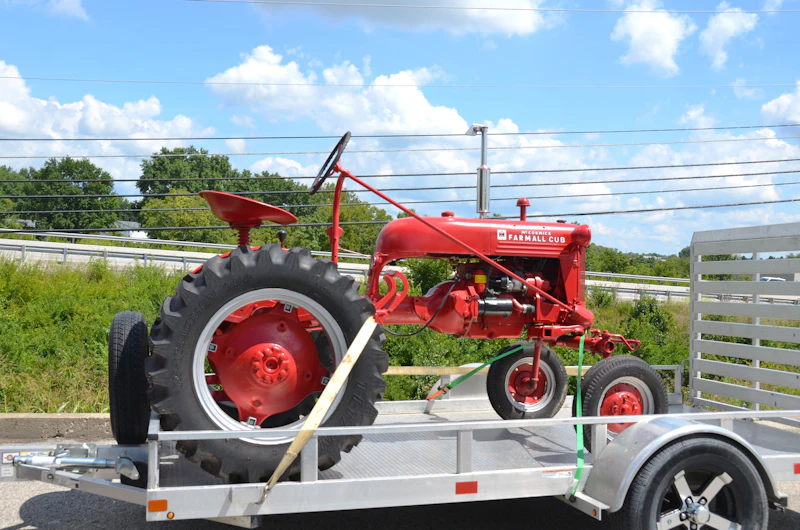 A small red vintage tractor held by a yellow strap on an aluminium flatbed trailer