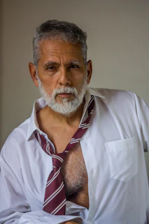 An older man with a grey beard in an unbuttoned white shirt and tie