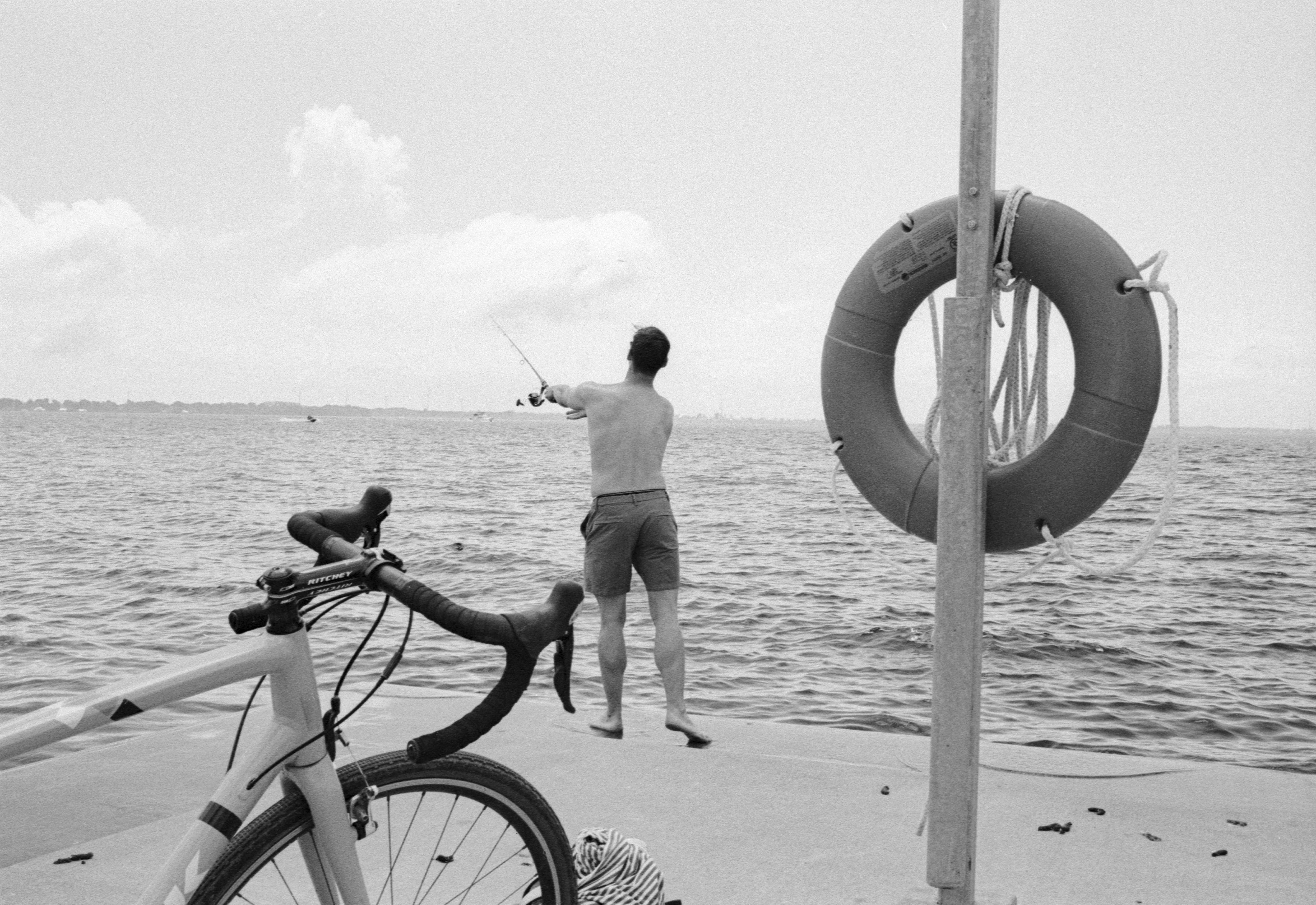 A shirtless man fishing from a pier with a bicycle and lifebuoy nearby