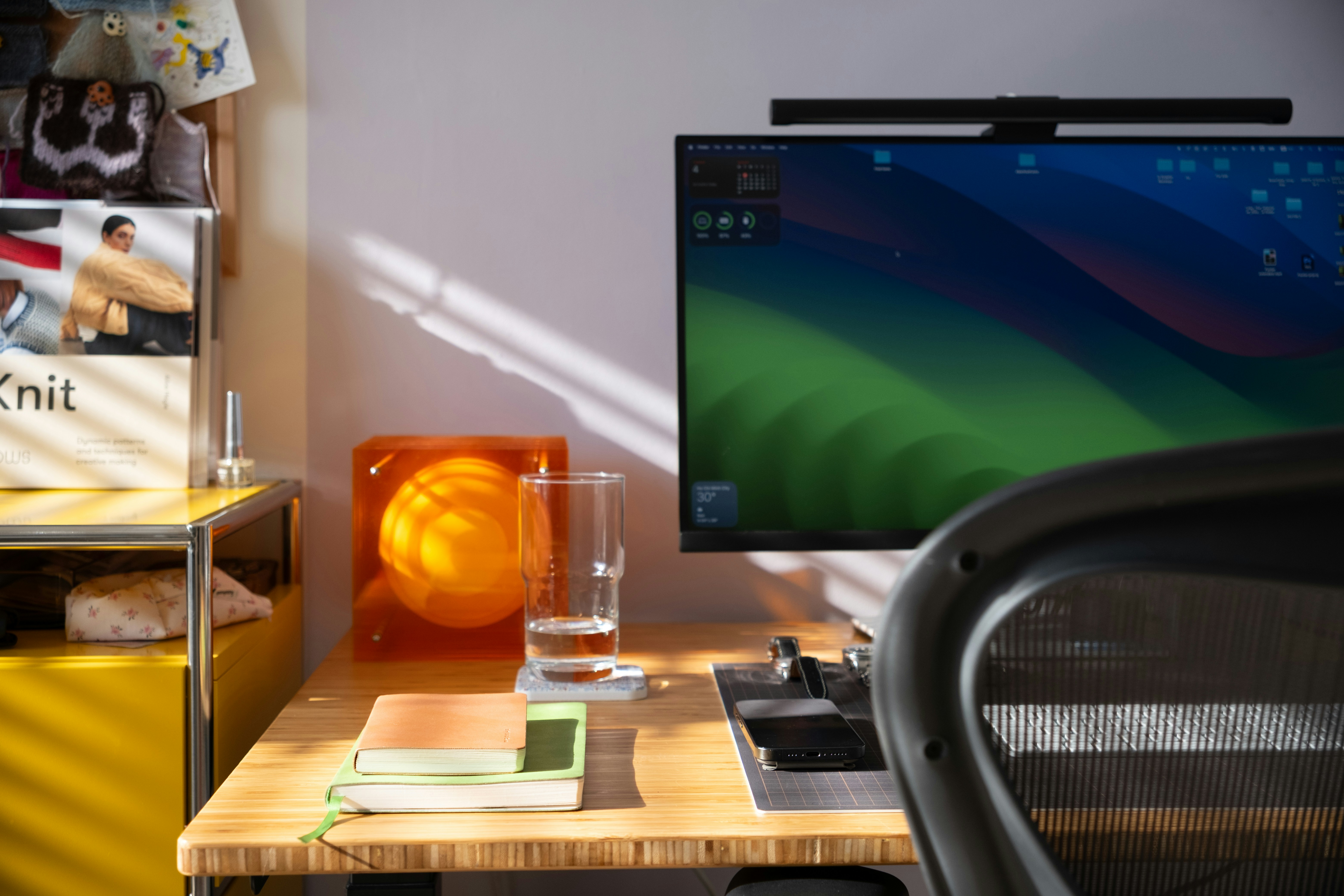 A wooden desk with a computer monitor, notebooks, and an orange acrylic lamp