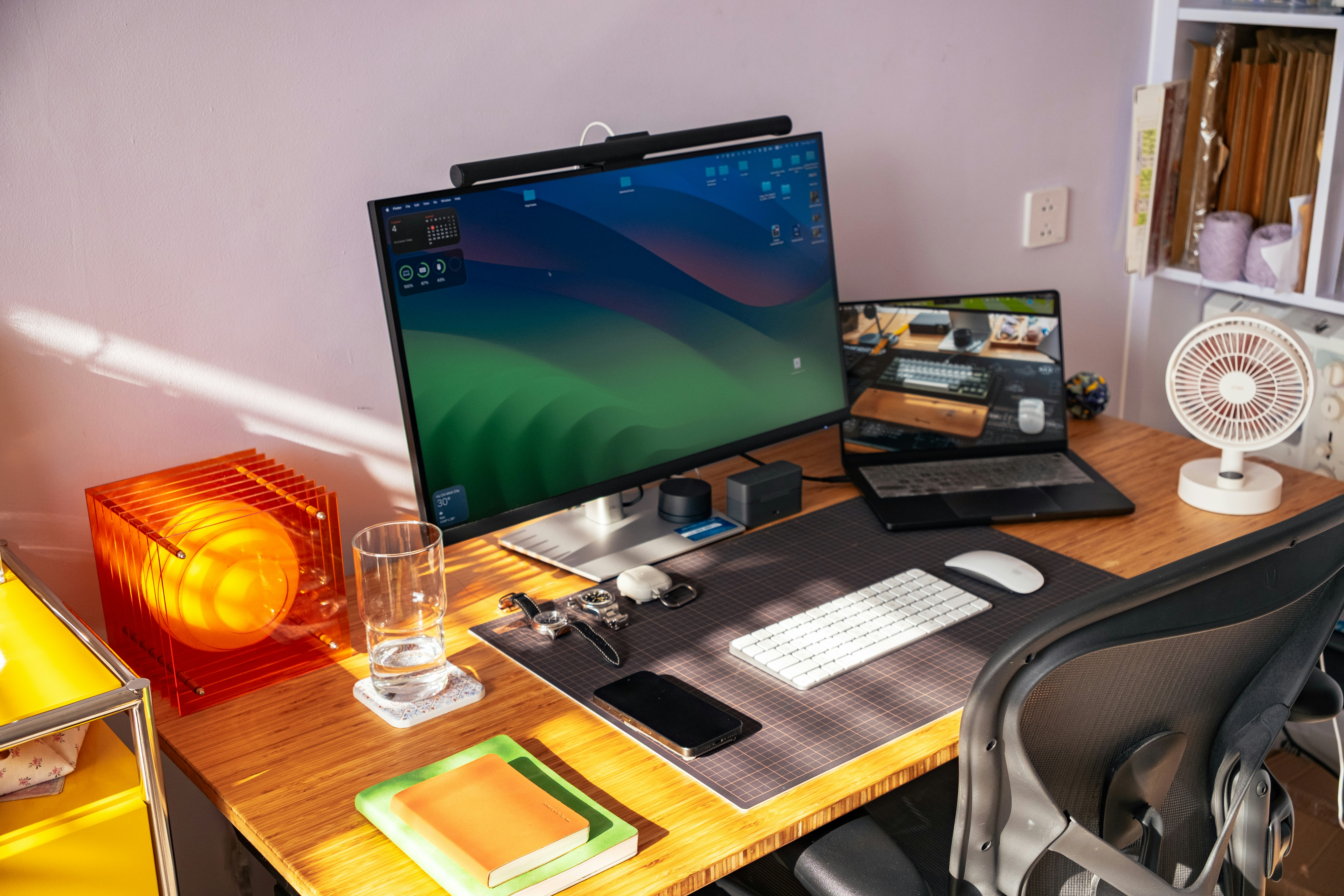 A computer monitor and laptop on a wooden desk in a sunlit workspace