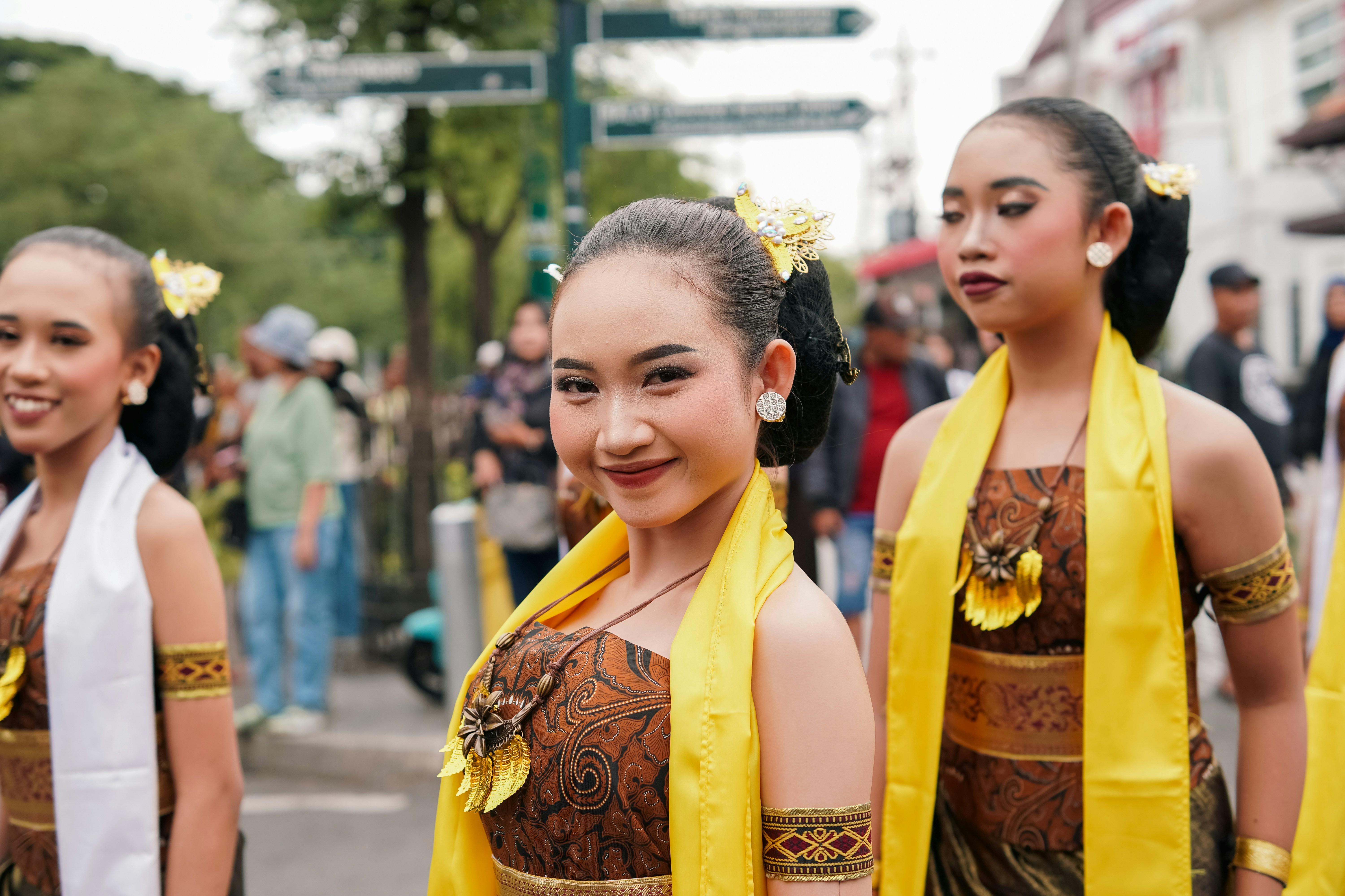 Three young women in traditional batik dresses and yellow sashes at a street parade