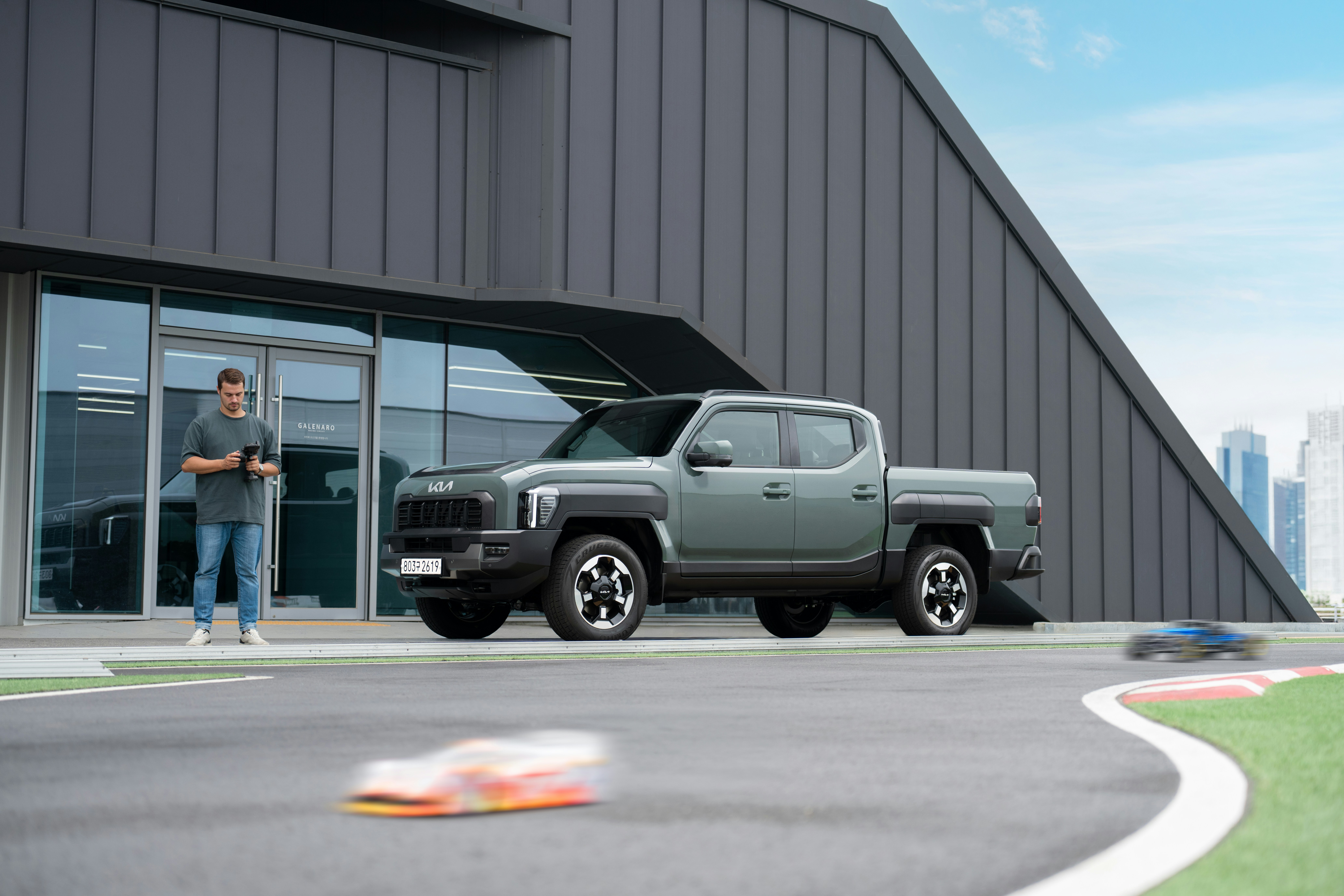 A man operating a remote control near a green Kia pickup truck