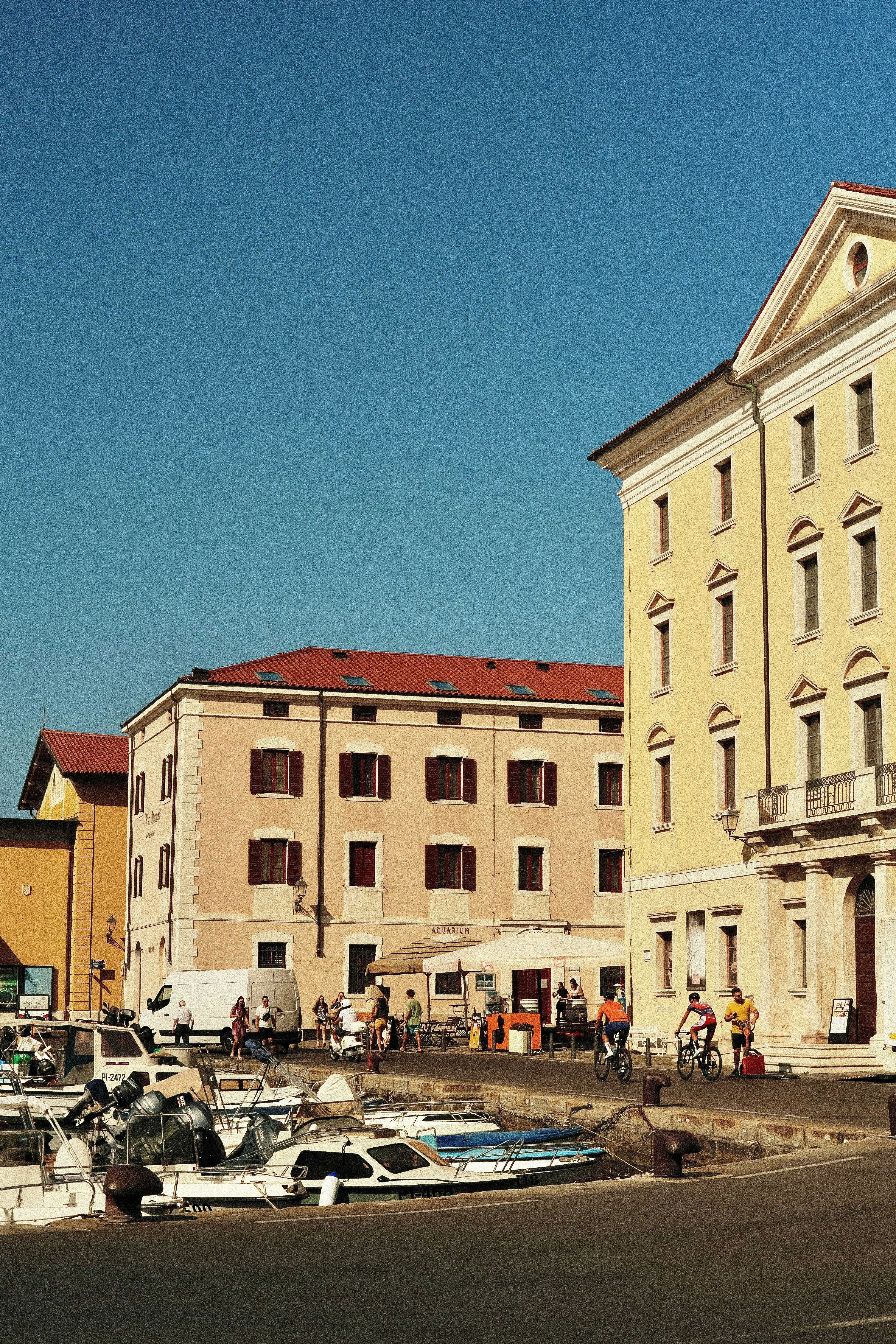 People cycling past a yellow building and boats moored at a harbor