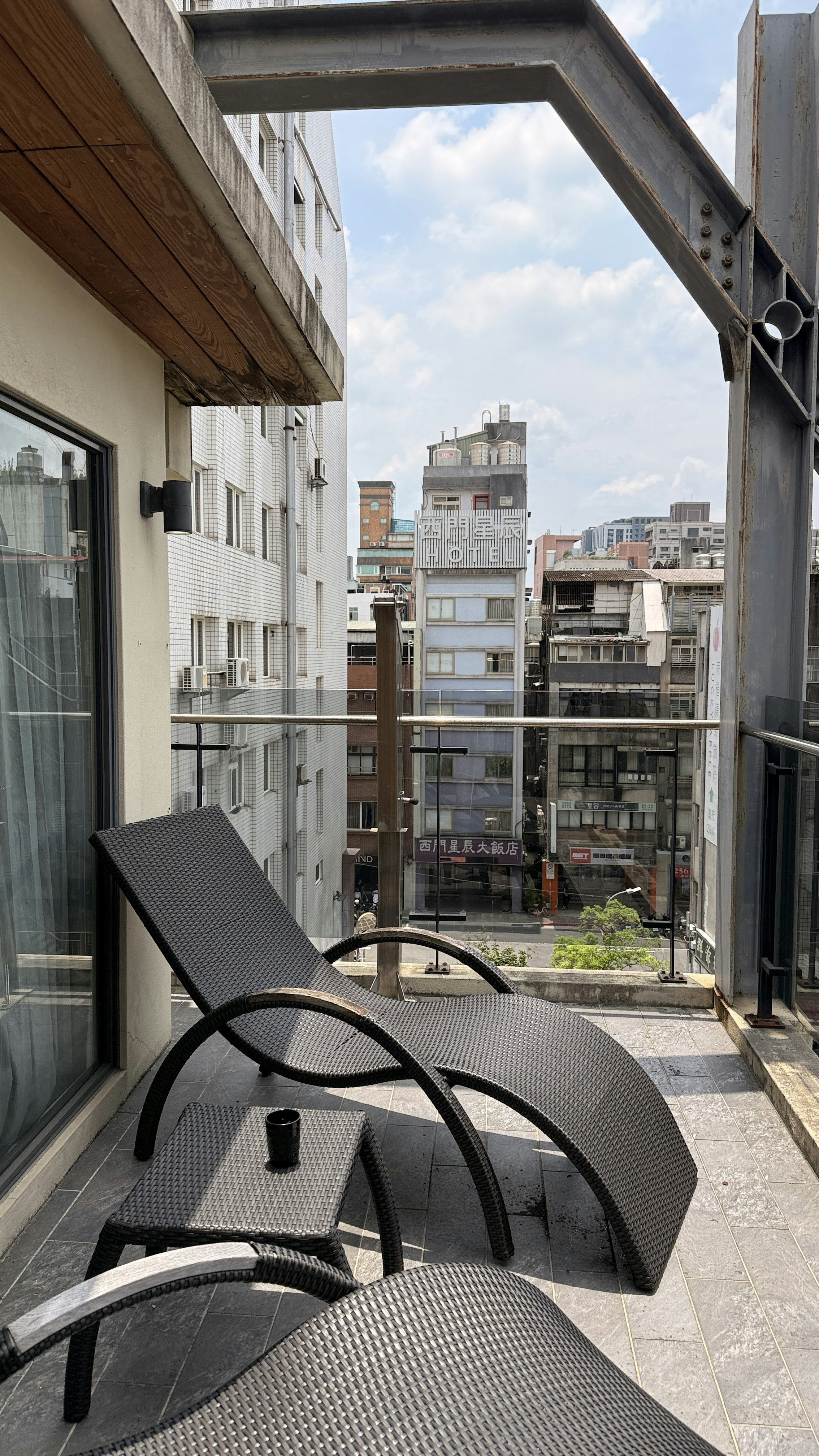 A black wicker lounge chair and small table on a balcony overlooking city buildings