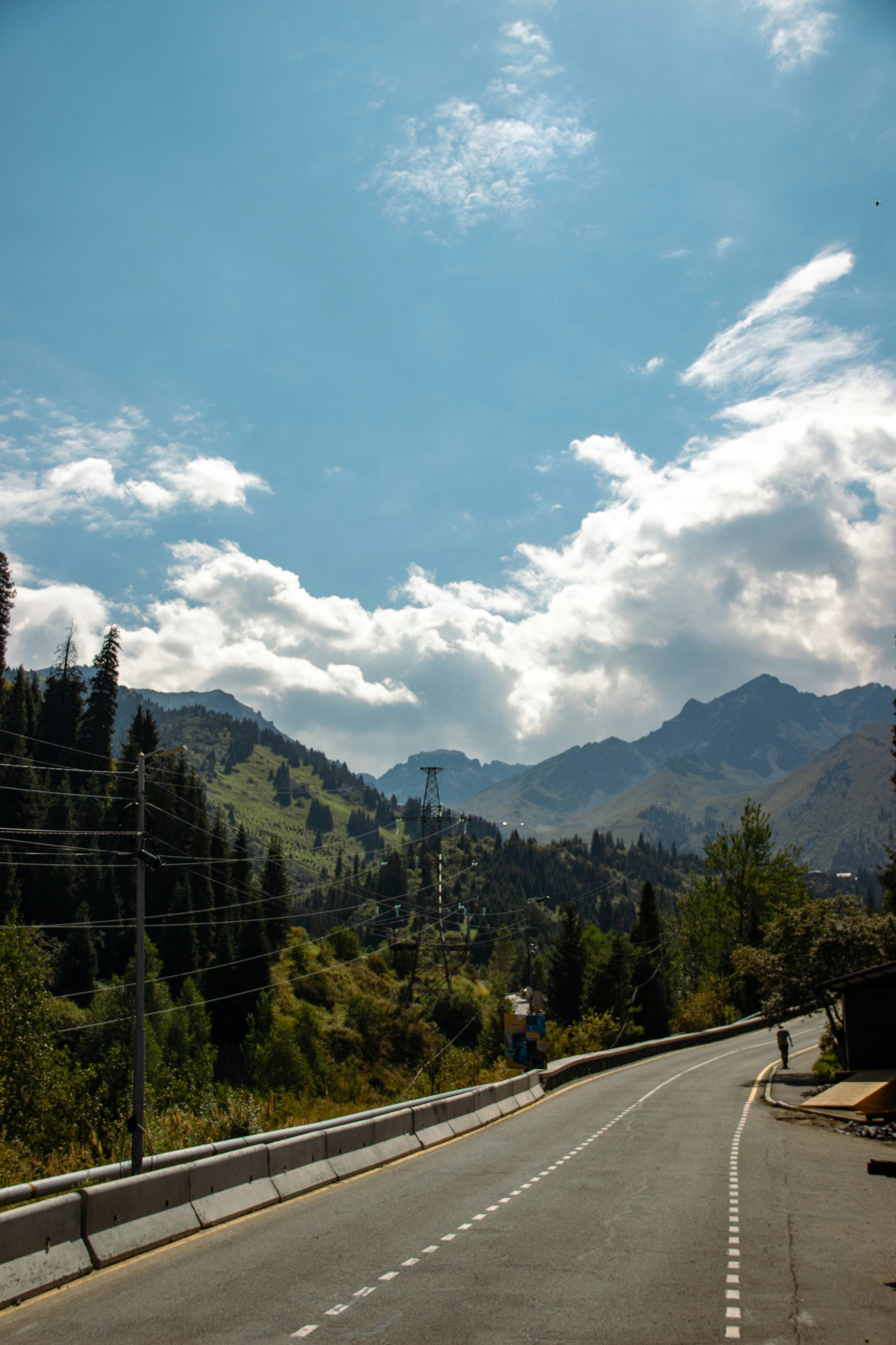 A winding asphalt road through a mountain valley with pine trees and clouds