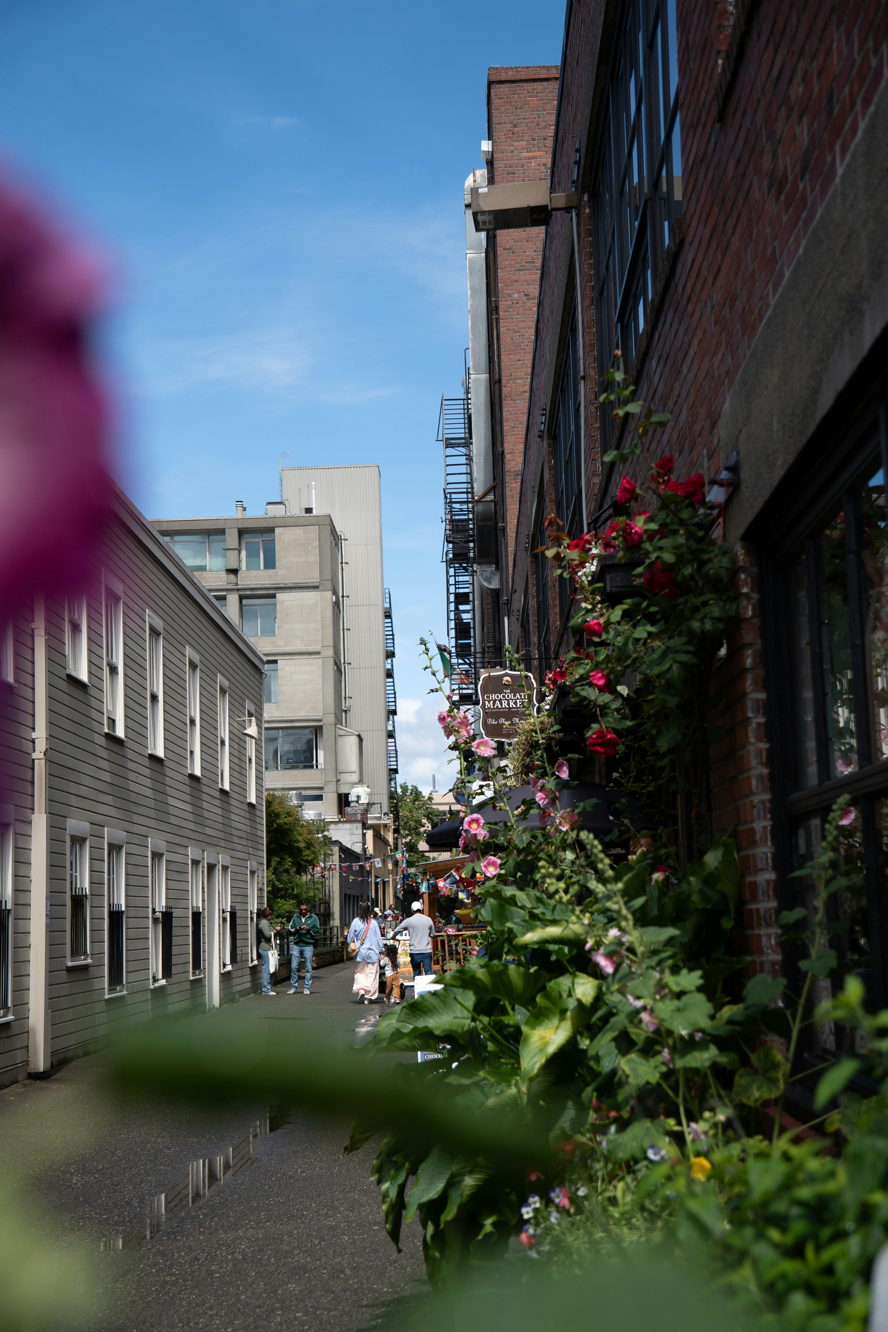 People walking down a narrow alleyway between brick and wooden buildings with flowers
