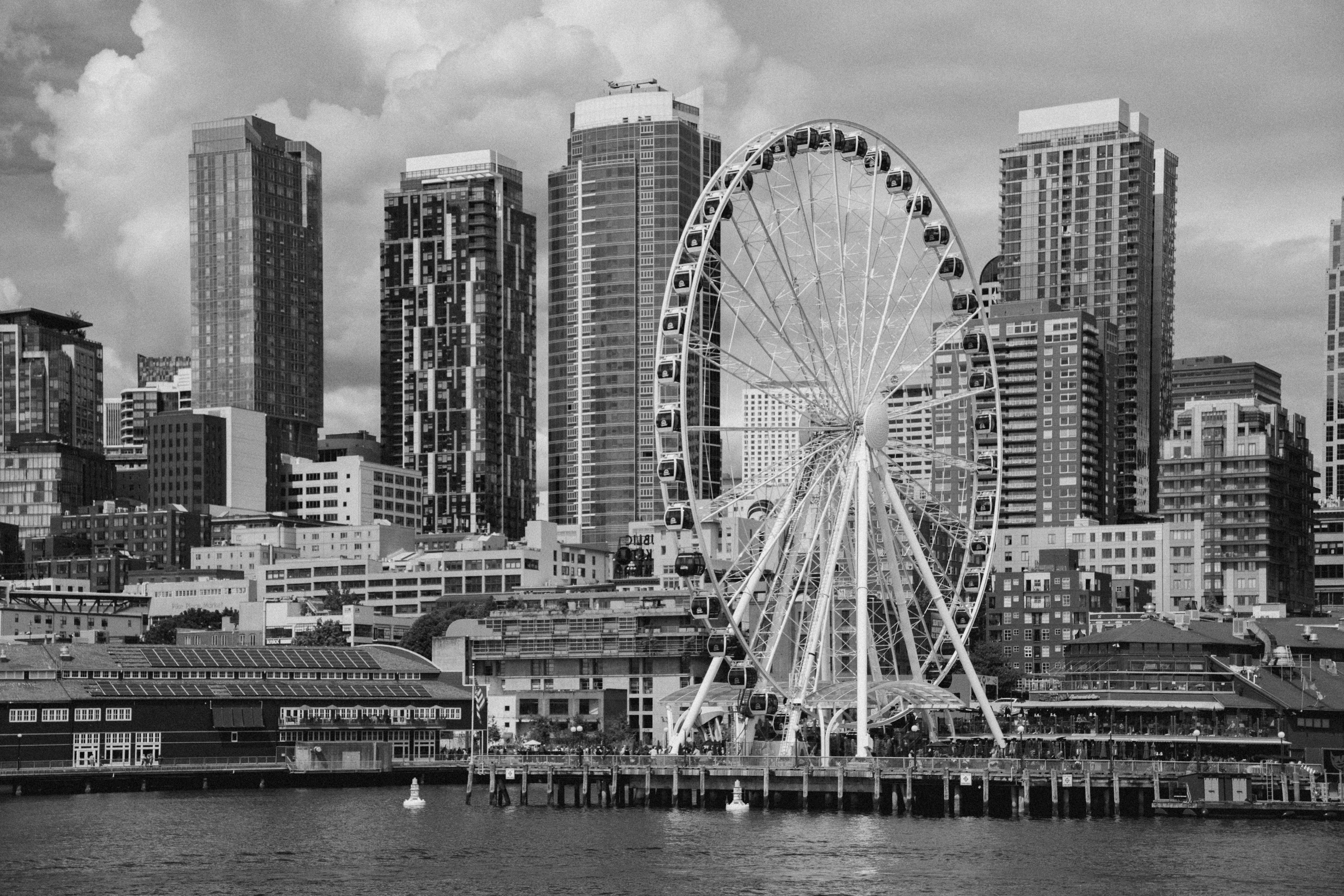 A large ferris wheel on a pier before a city skyline in black and white