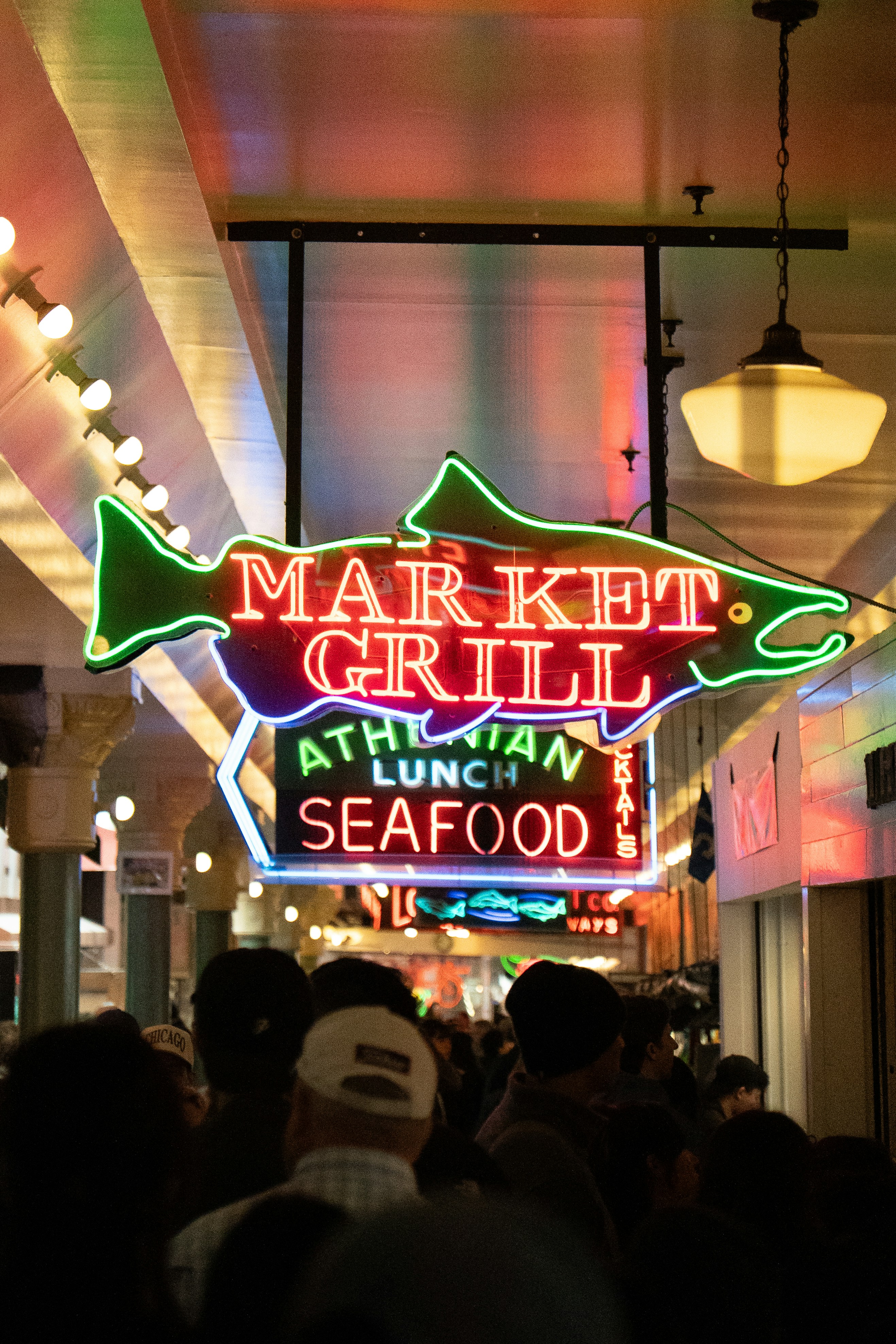 A crowd of people walking beneath a neon fish sign reading Market Grill