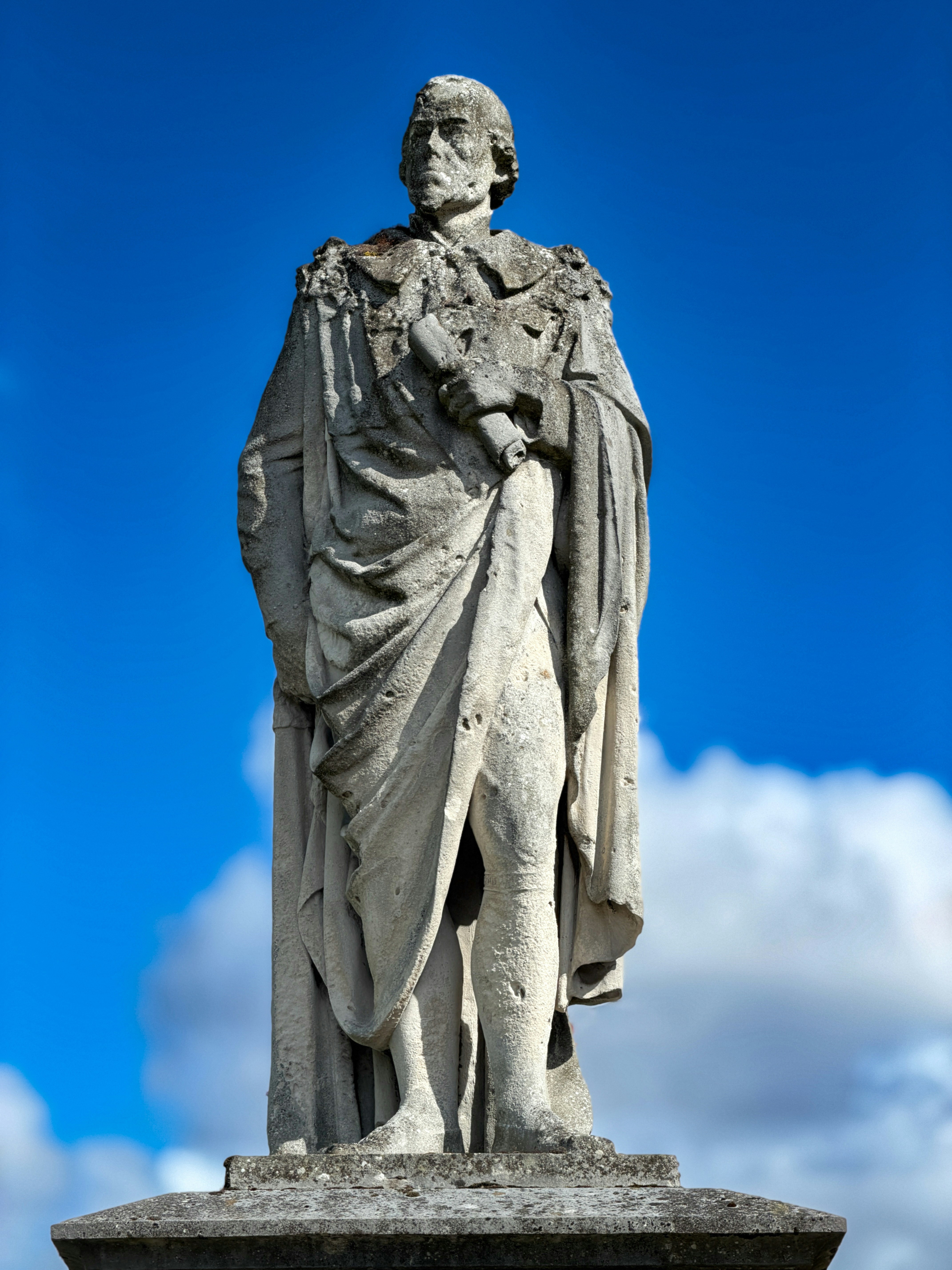 A weathered stone statue of a man in robes holding a scroll against blue sky