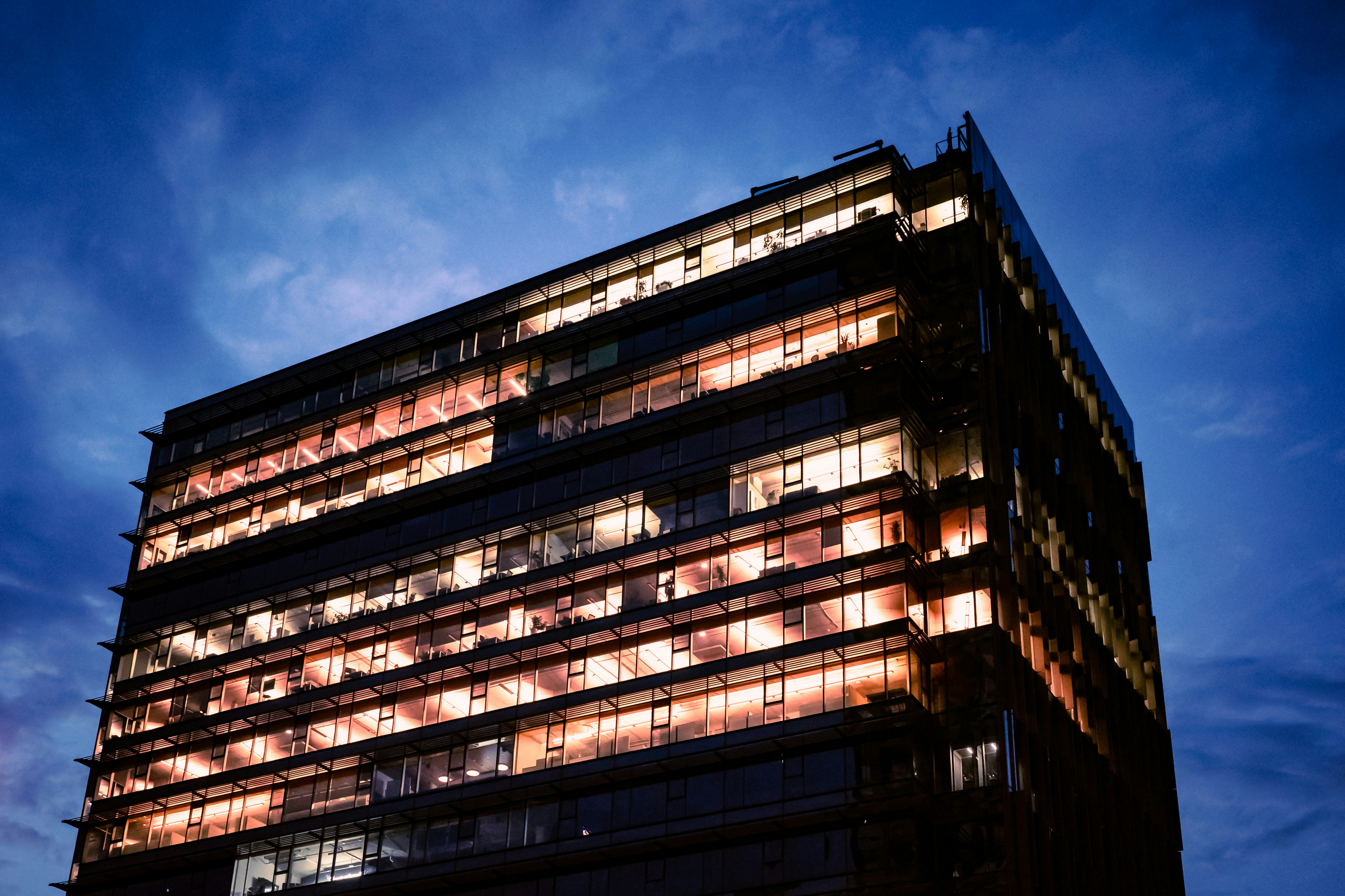 Lit windows glow in an office building under a deep blue evening sky