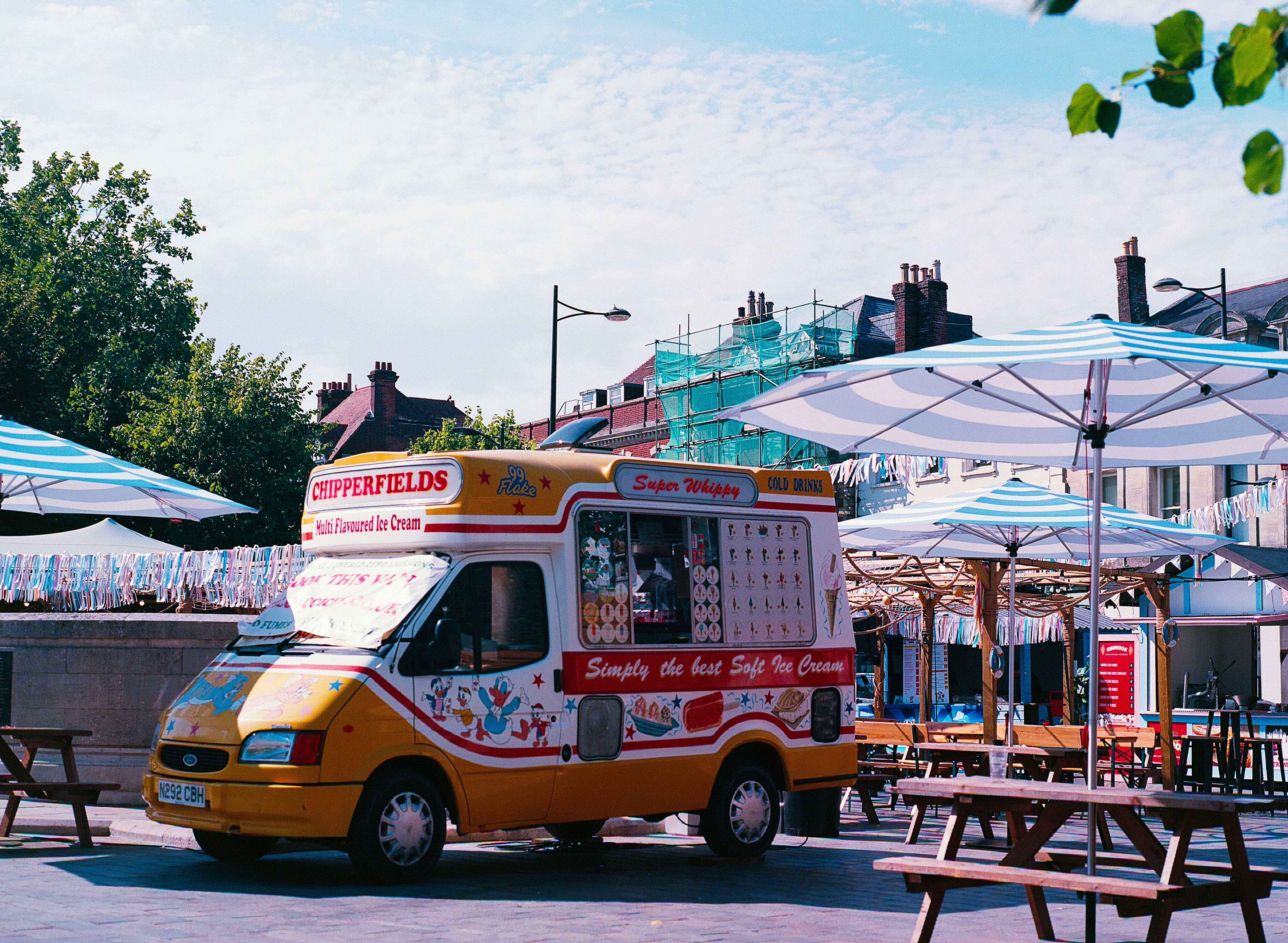 A yellow and white ice cream van parked in a sunny outdoor plaza