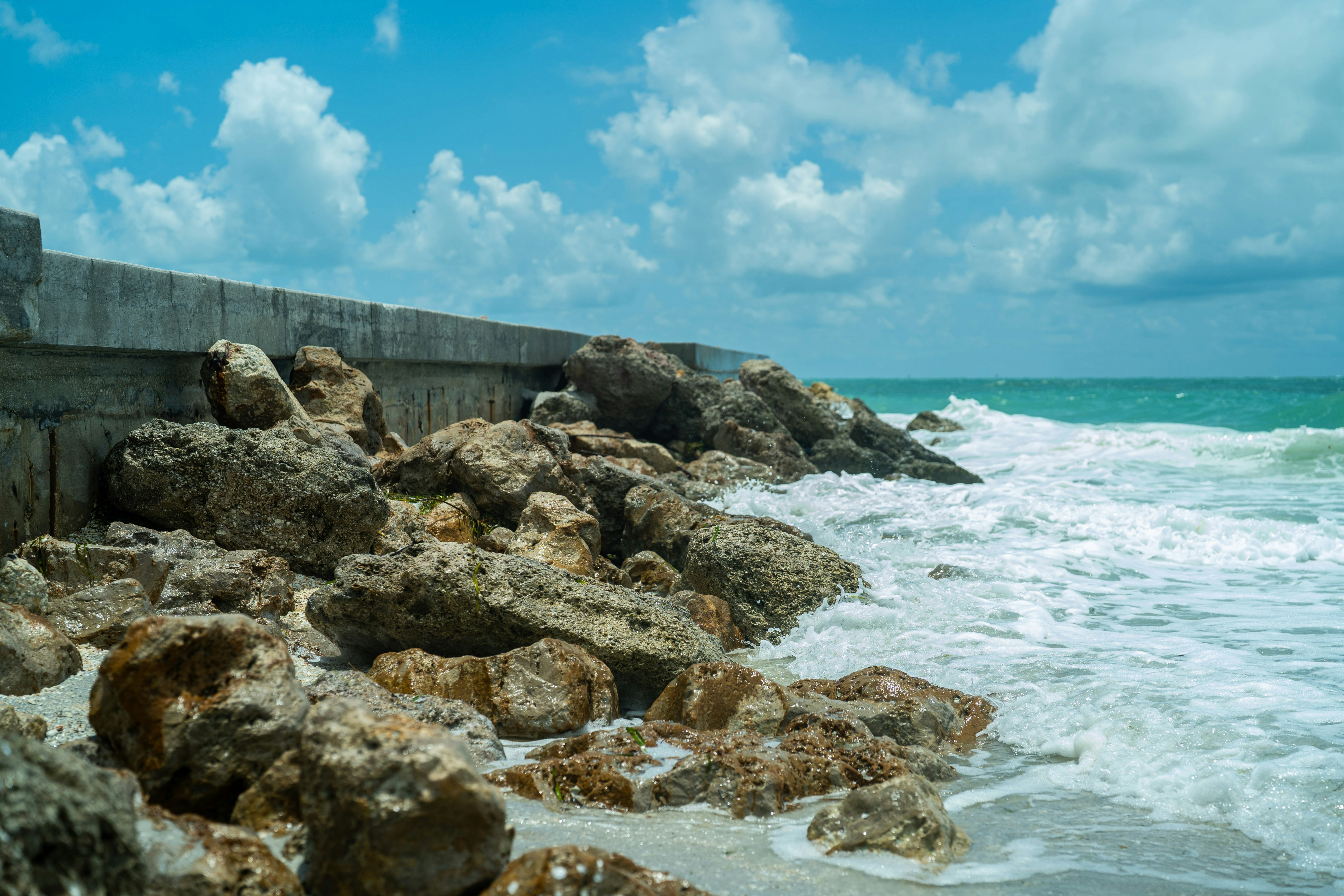 Waves crashing against a concrete seawall and large rocks on a sandy beach