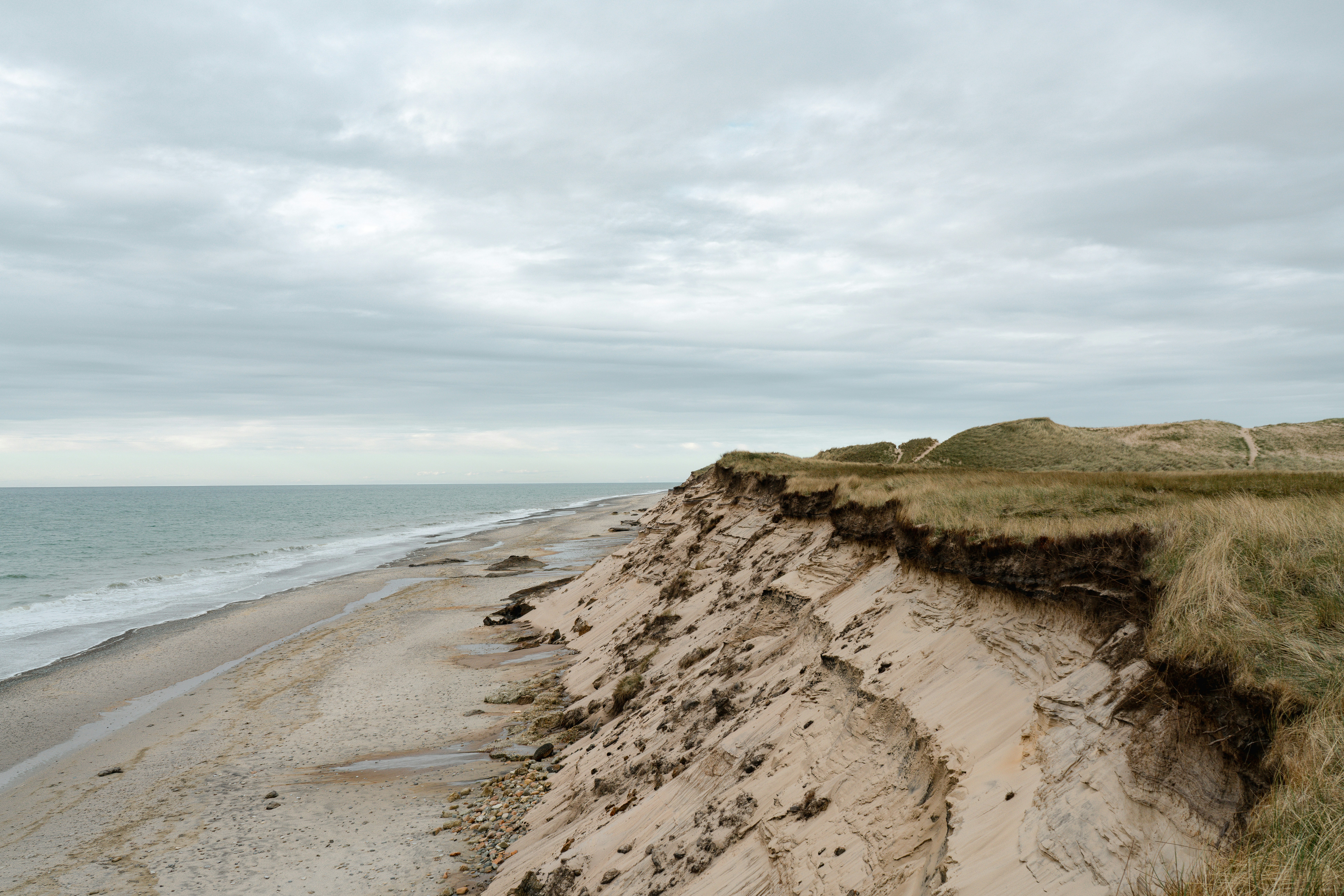 A sandy beach below a steep eroding cliff with grass under a cloudy sky