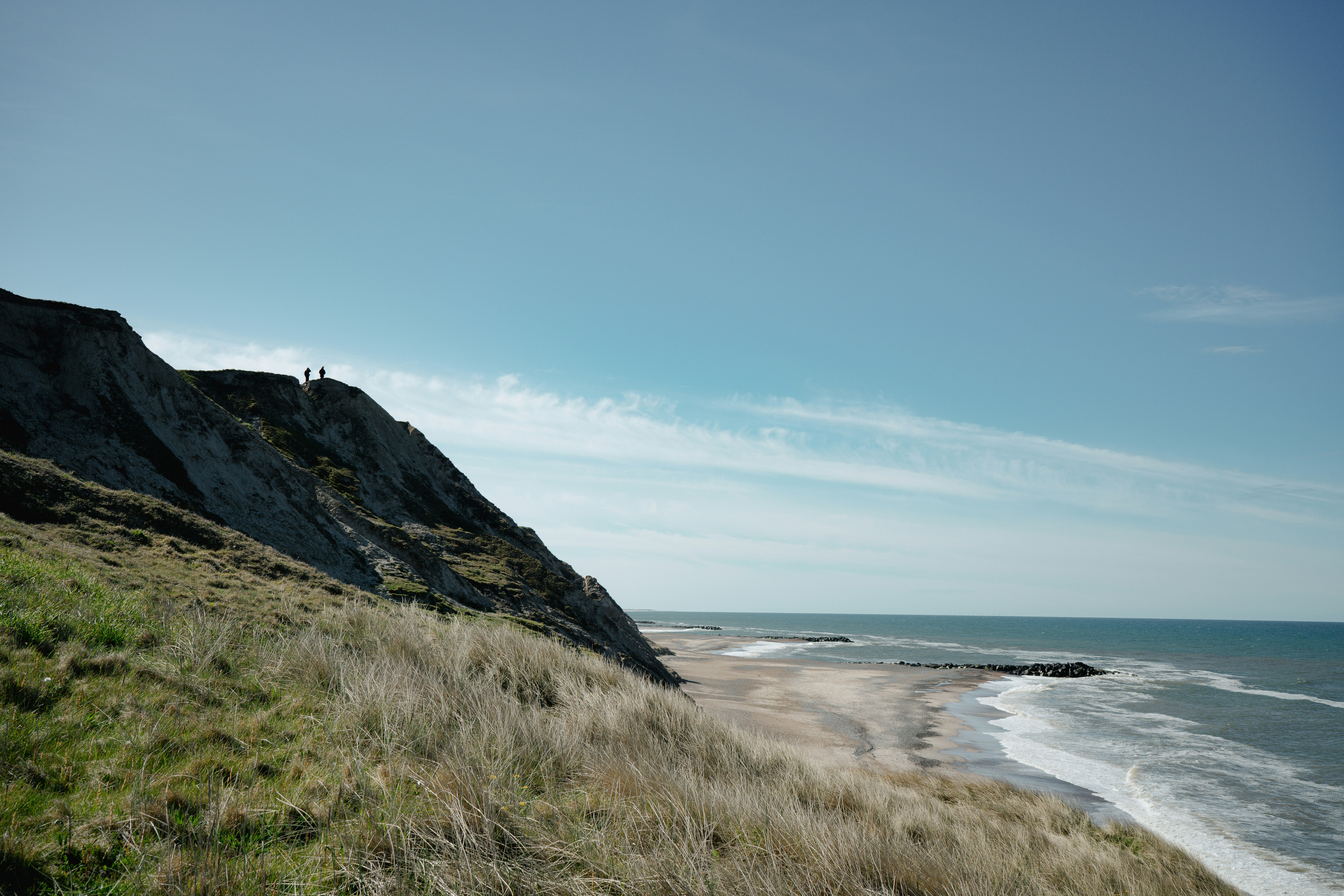 Two people on a grassy cliff overlooking a sandy beach and the ocean