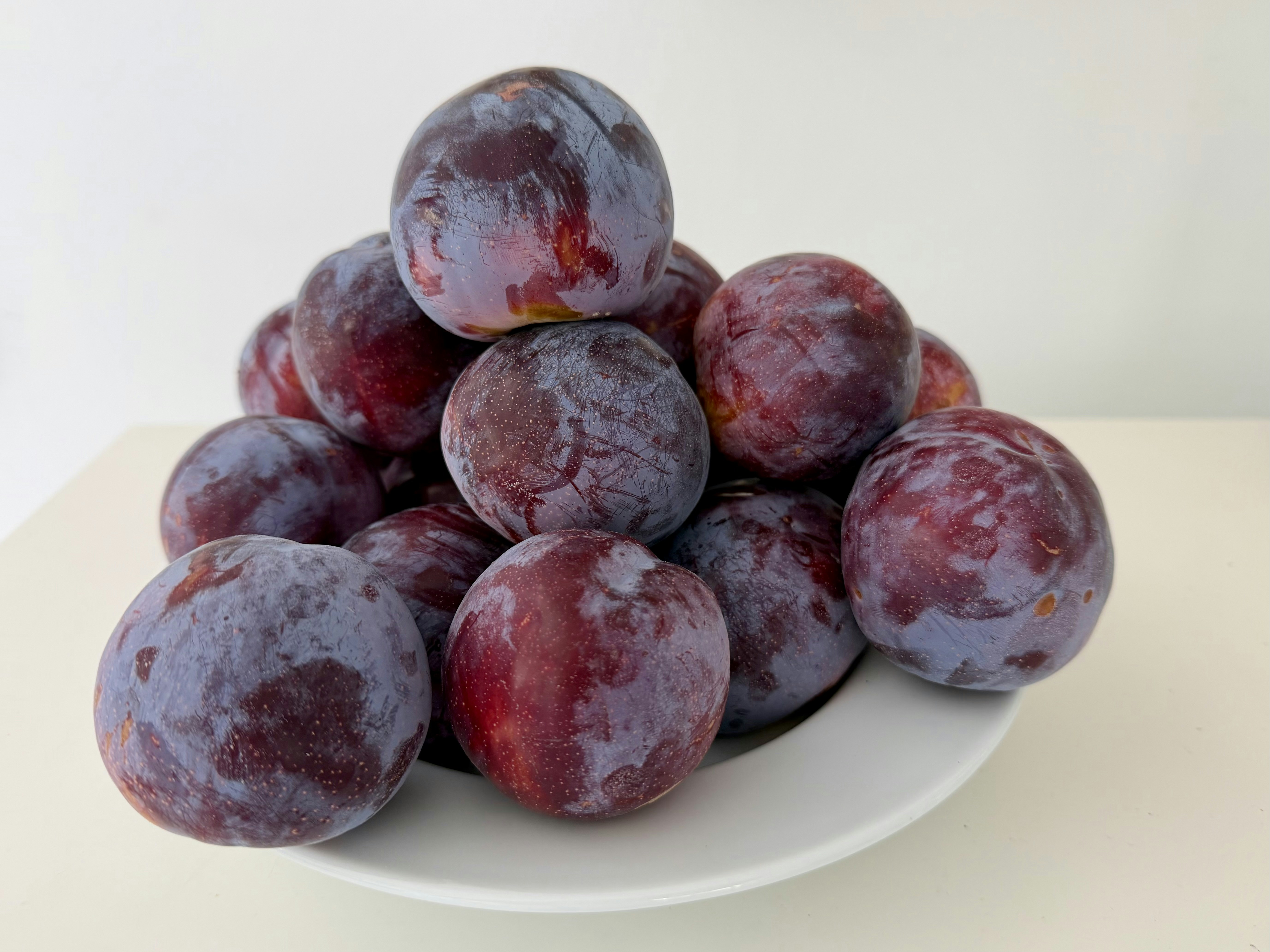 A pile of ripe purple plums in a white bowl on a white surface
