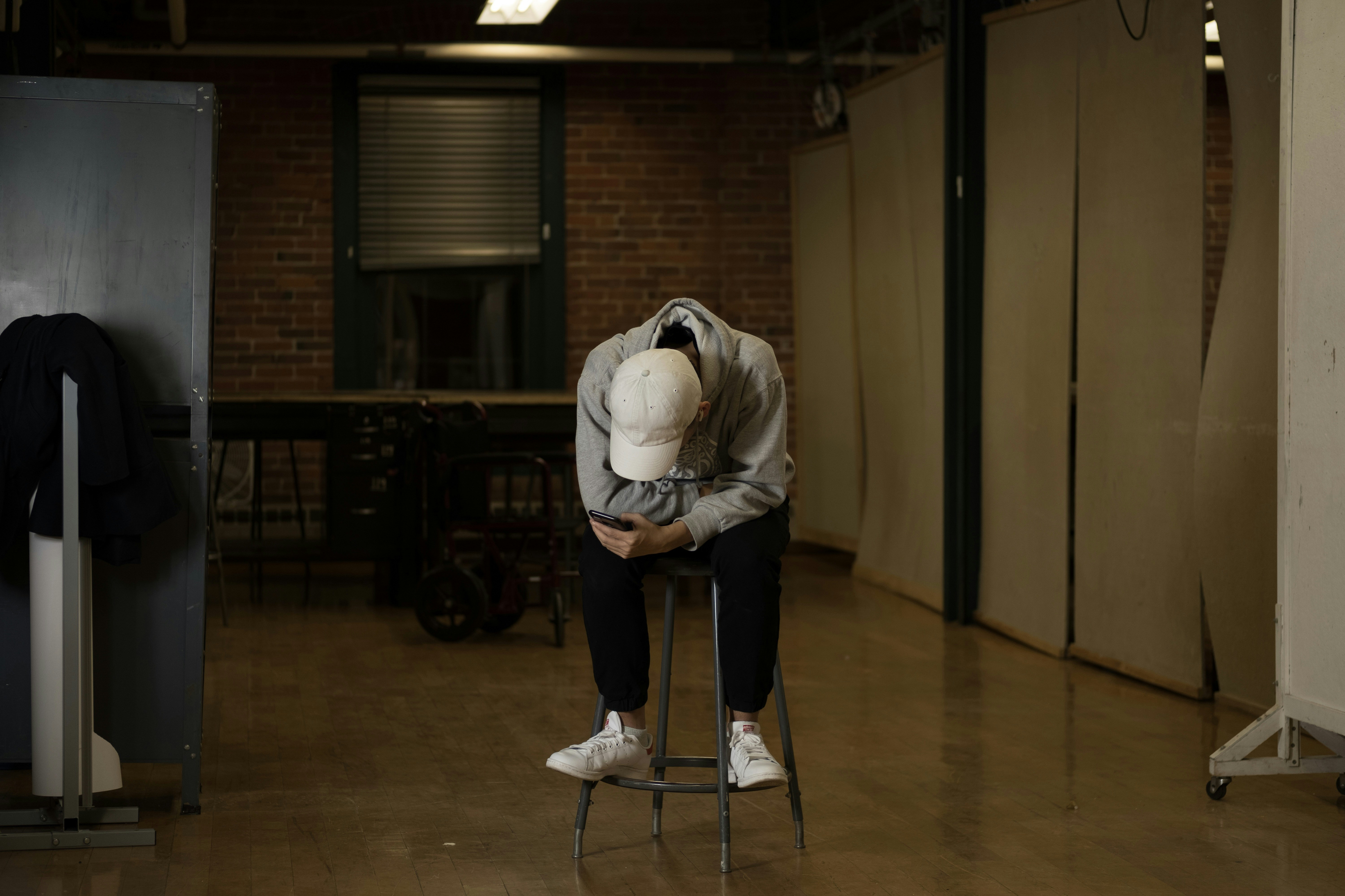 A person in a grey hoodie and white cap using a phone on a stool