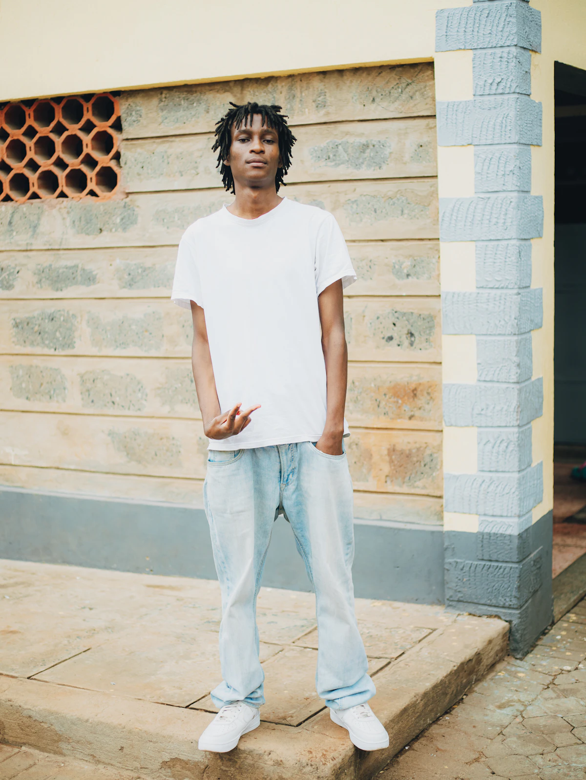 Rapaz de camiseta branca lisa, calça jeans clara e tênis branco encostado em uma parede