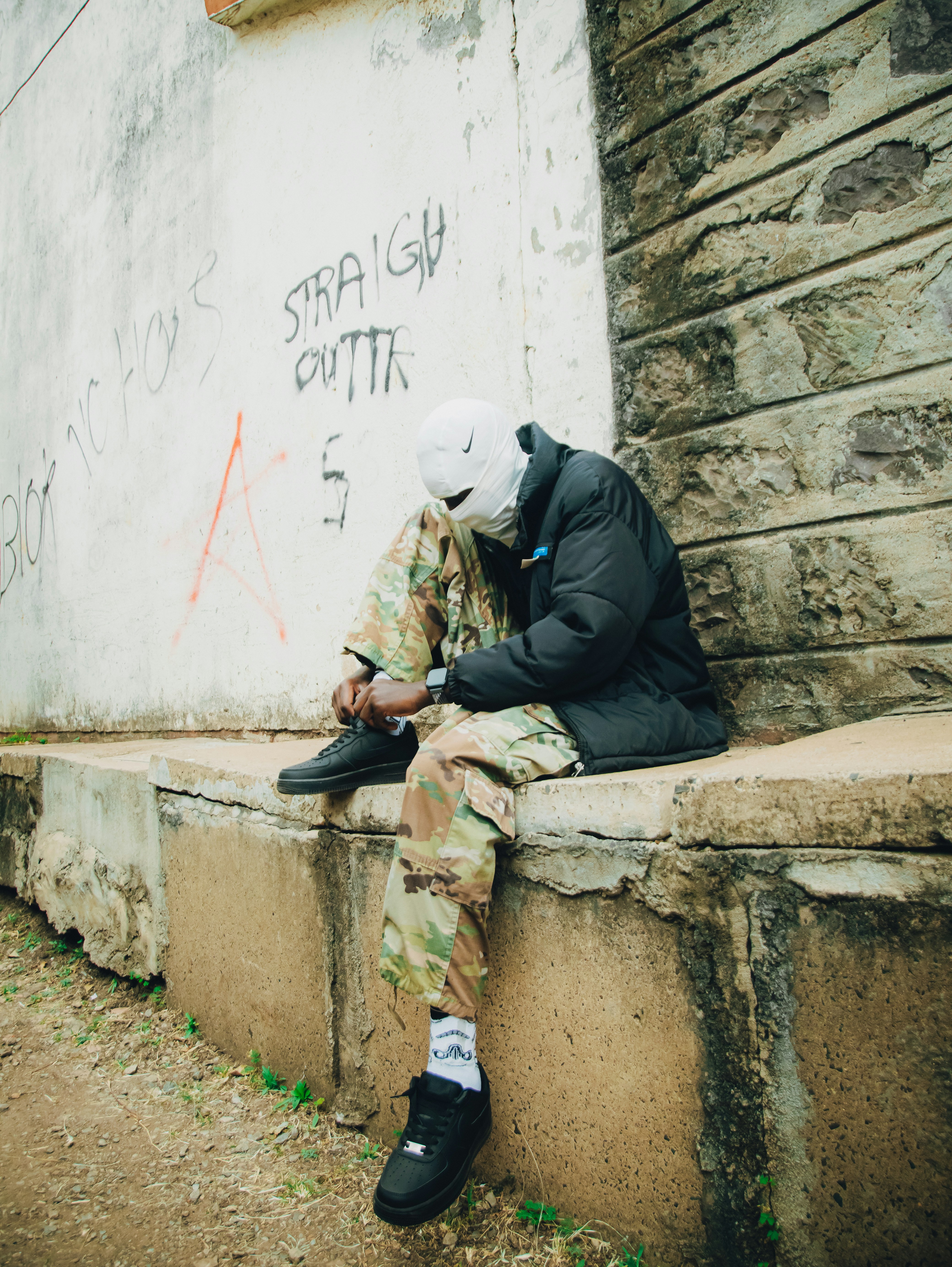 A person in a white balaclava and camouflage pants tying black sneakers on a ledge