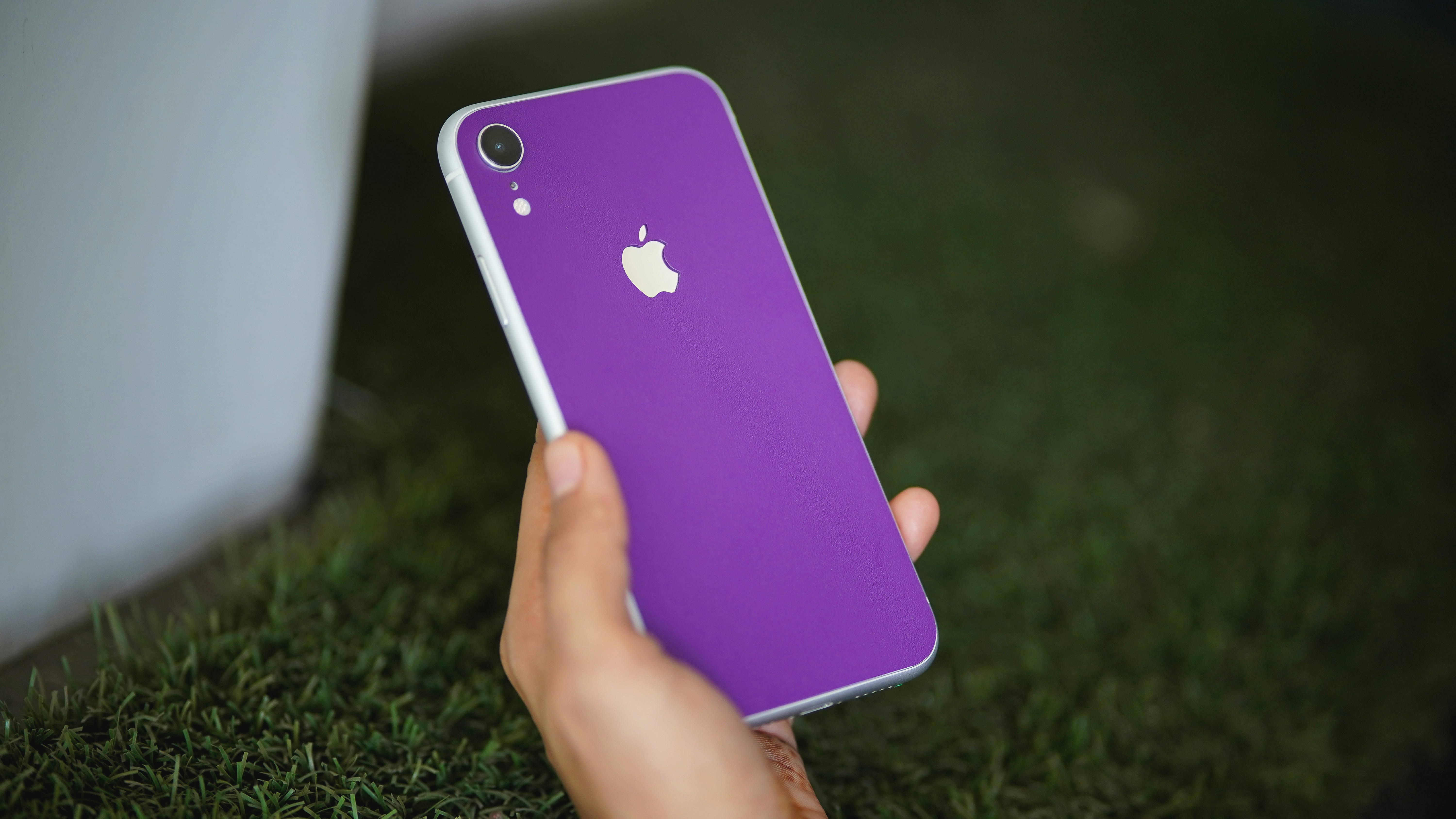 A hand holding a purple smartphone with a silver logo against green grass