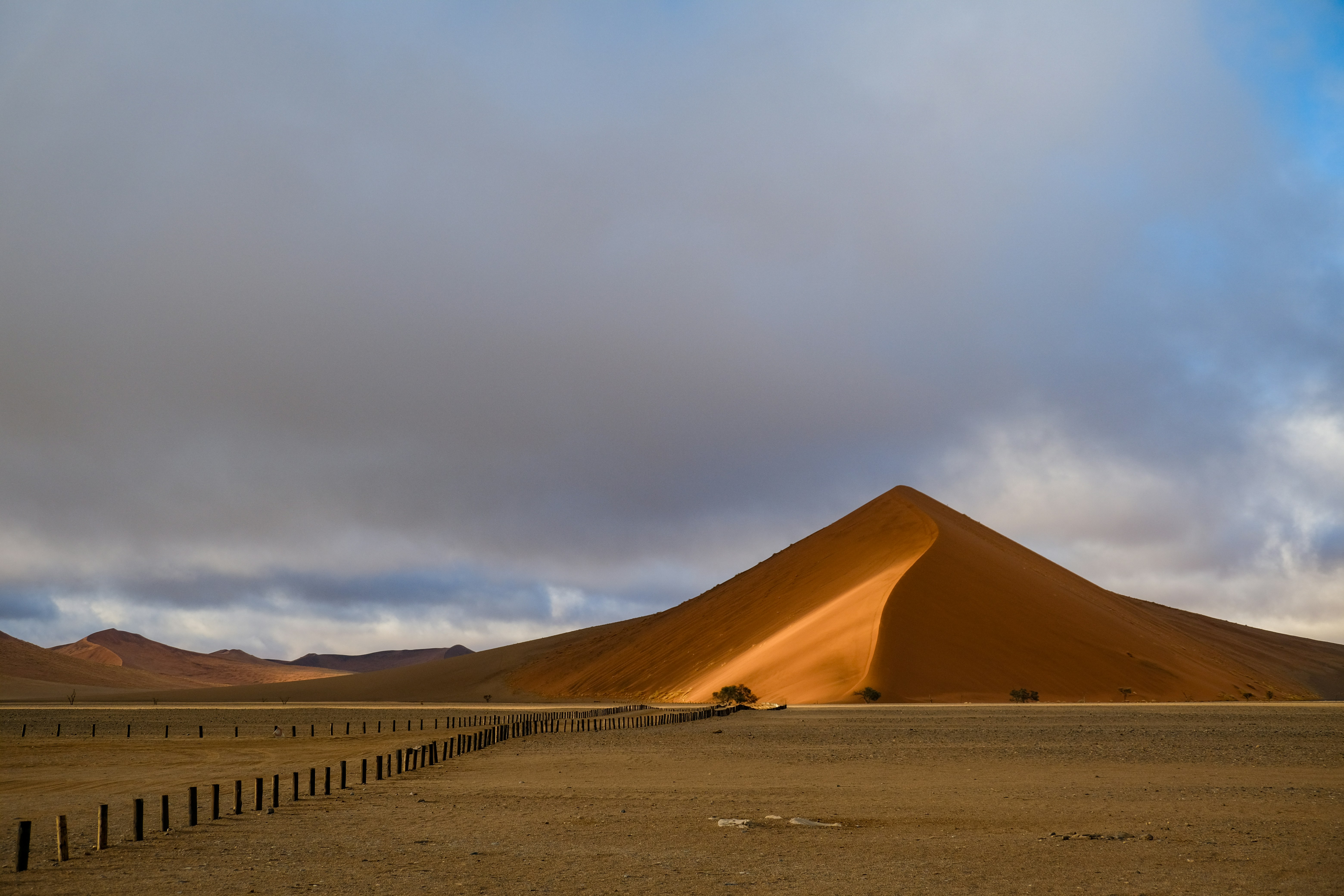 A large orange sand dune under a cloudy sky with a wooden fence
