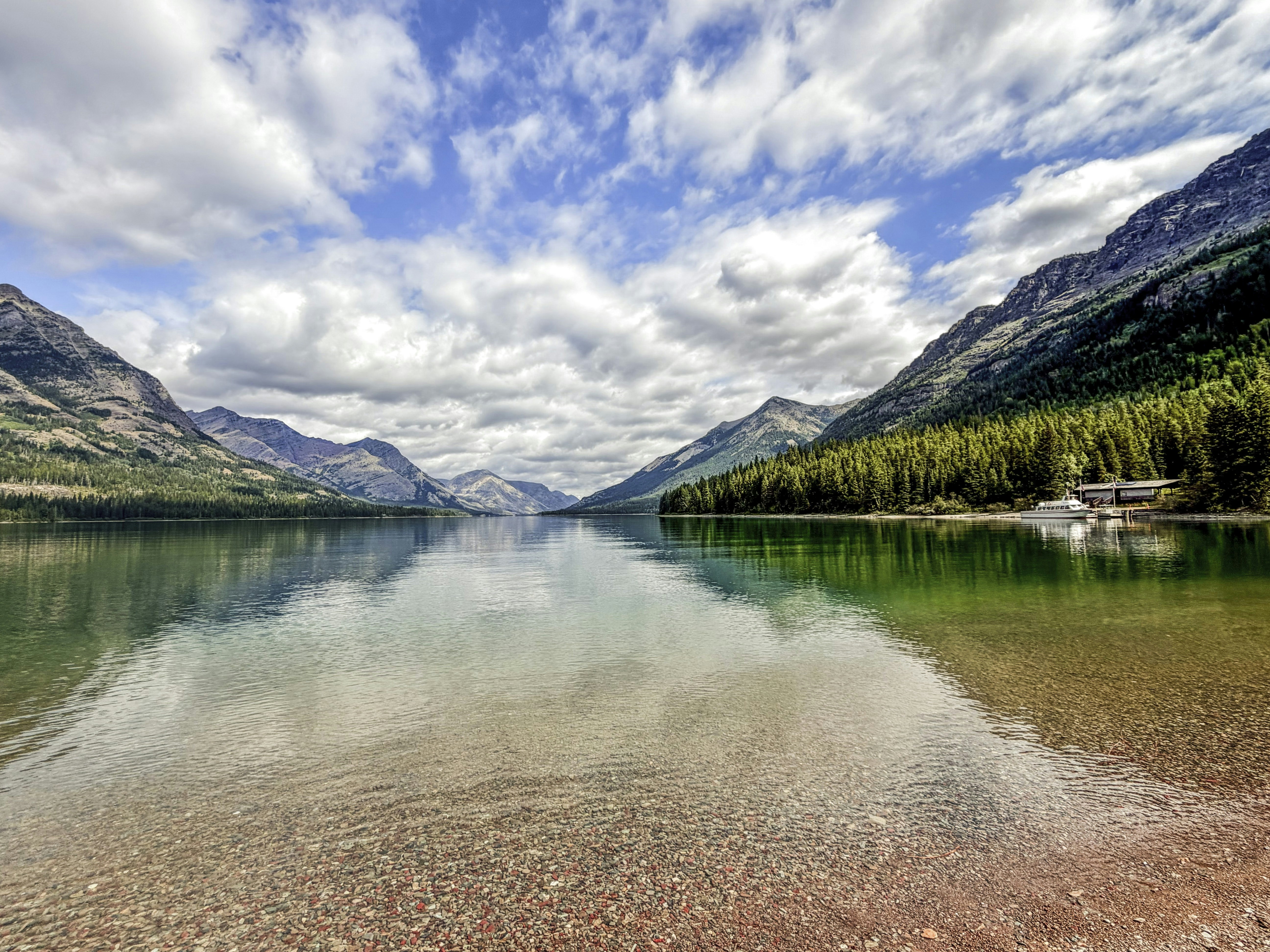 A clear lake reflecting mountains and a cloudy blue sky with a small boat