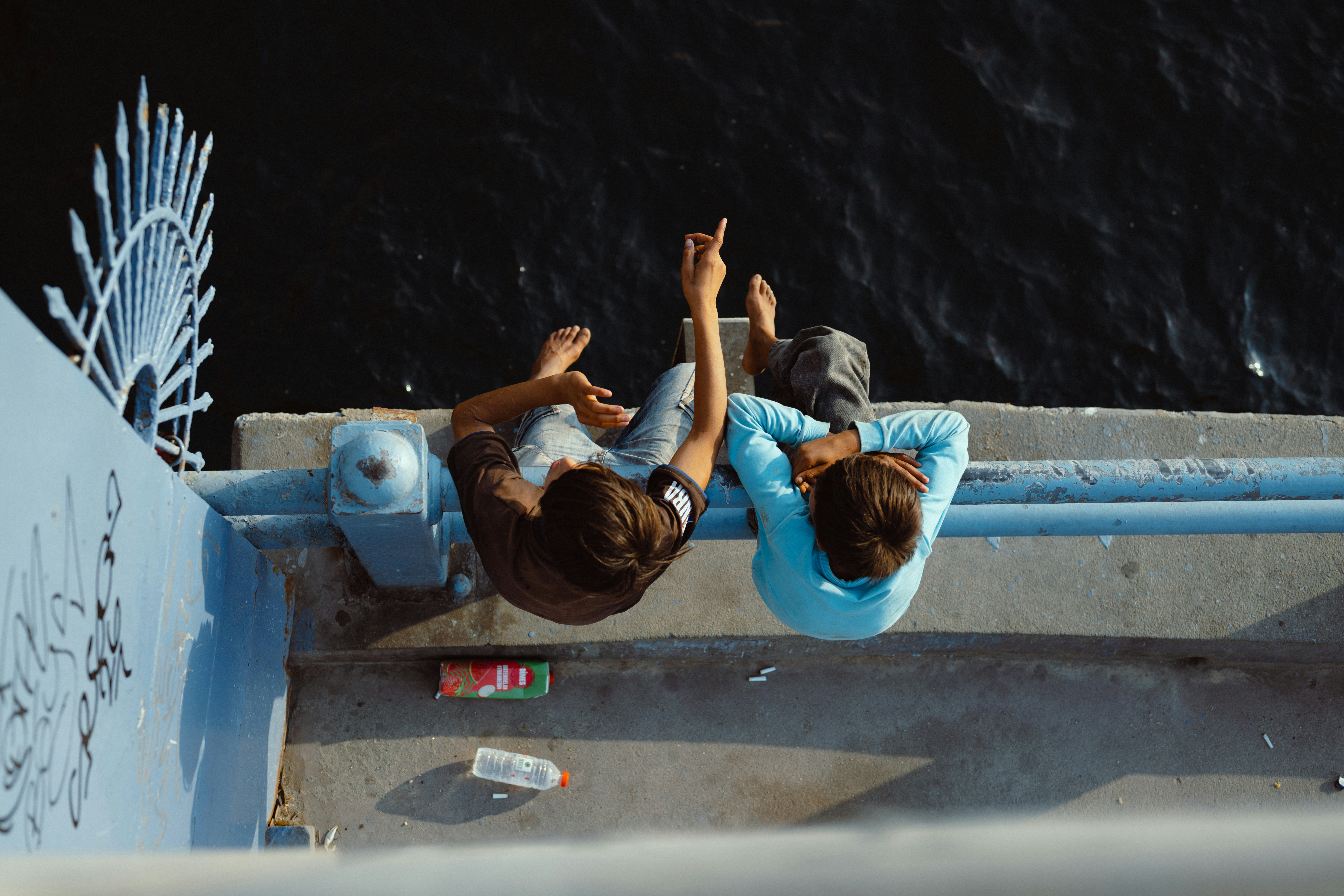 Two people sitting on a blue bridge railing pointing toward dark water below - 相关推荐