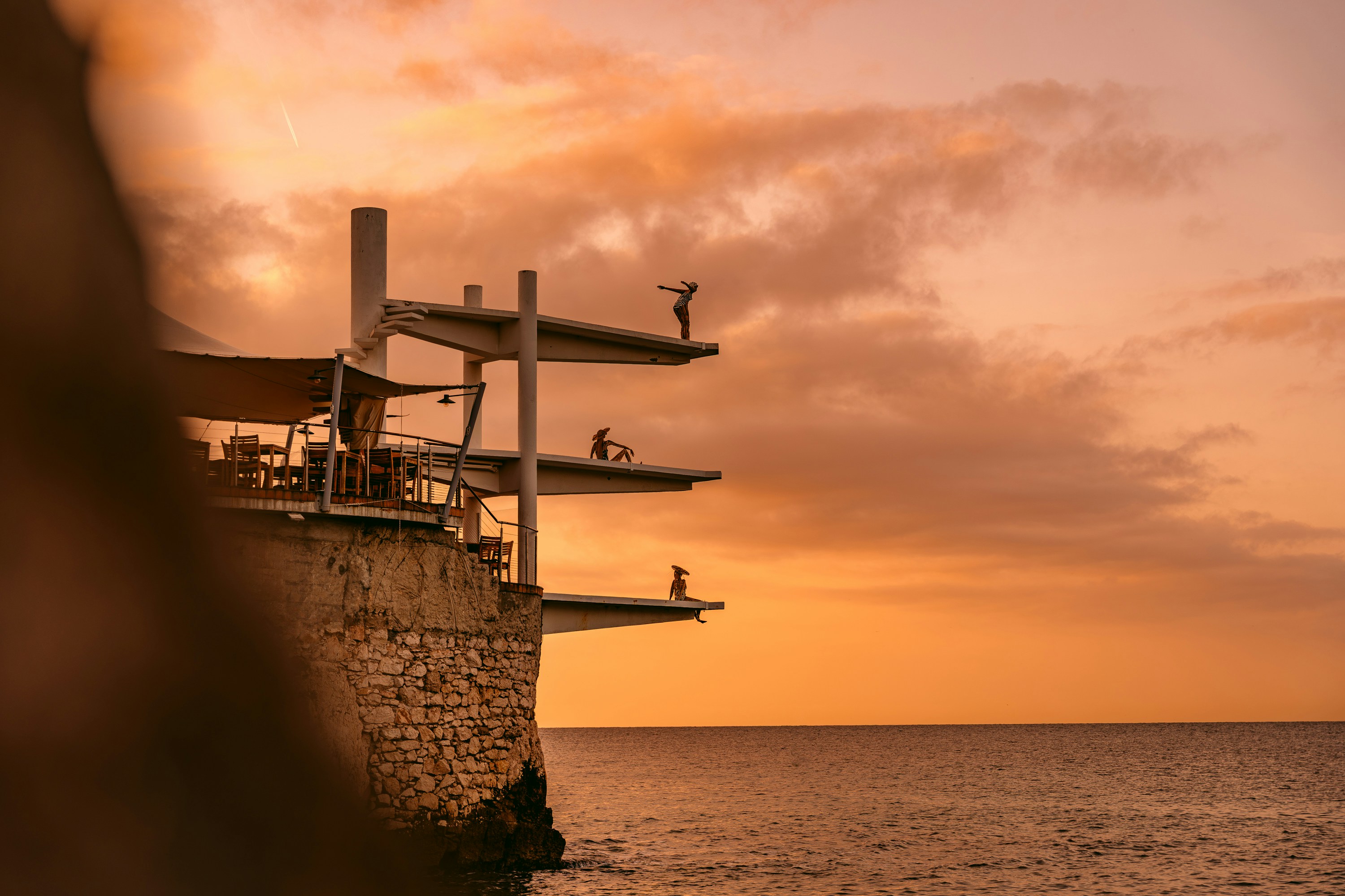 Three people on tiered diving platforms extending from a stone cliff at golden hour