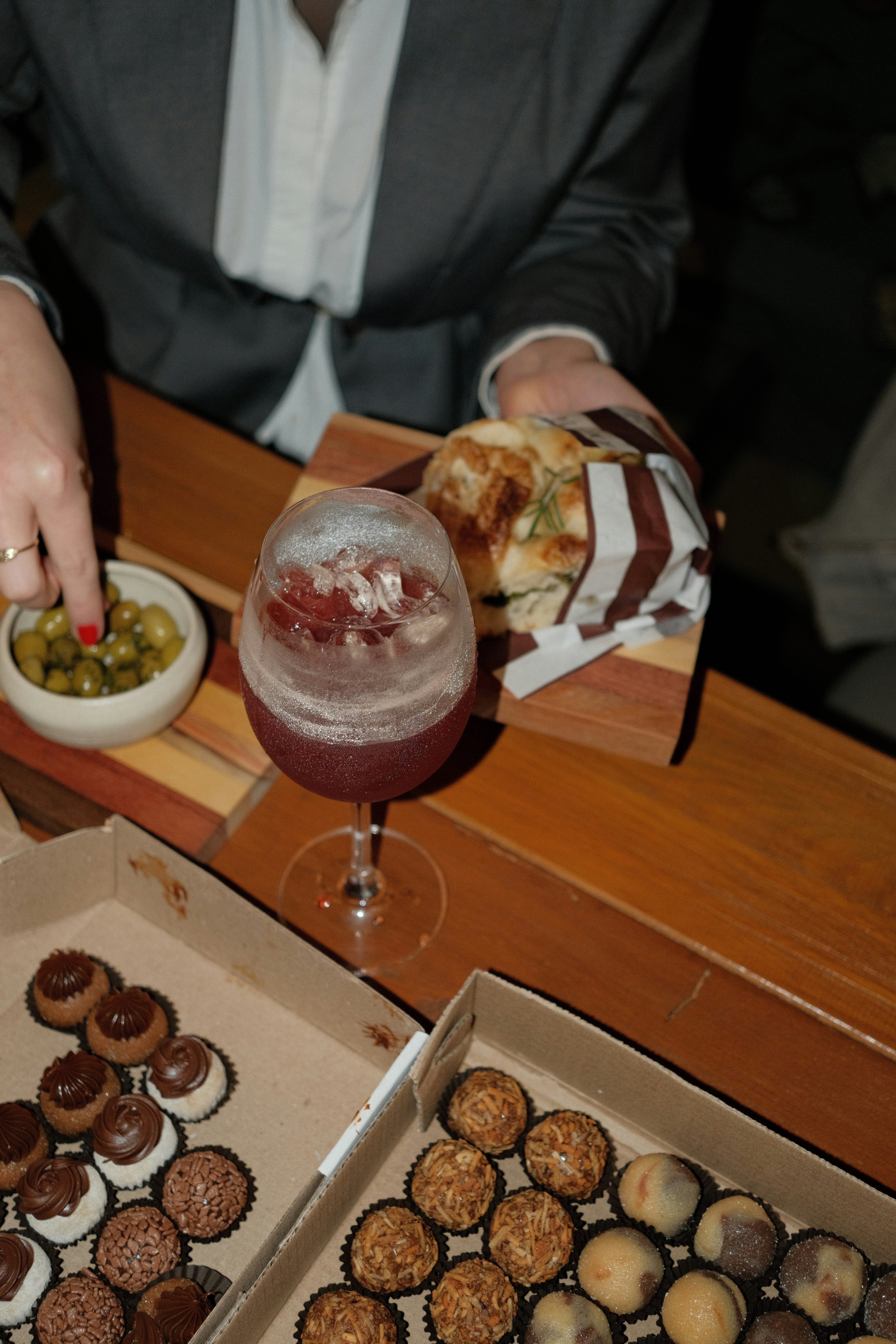 A person in a grey blazer reaching for olives beside a cocktail and pastries