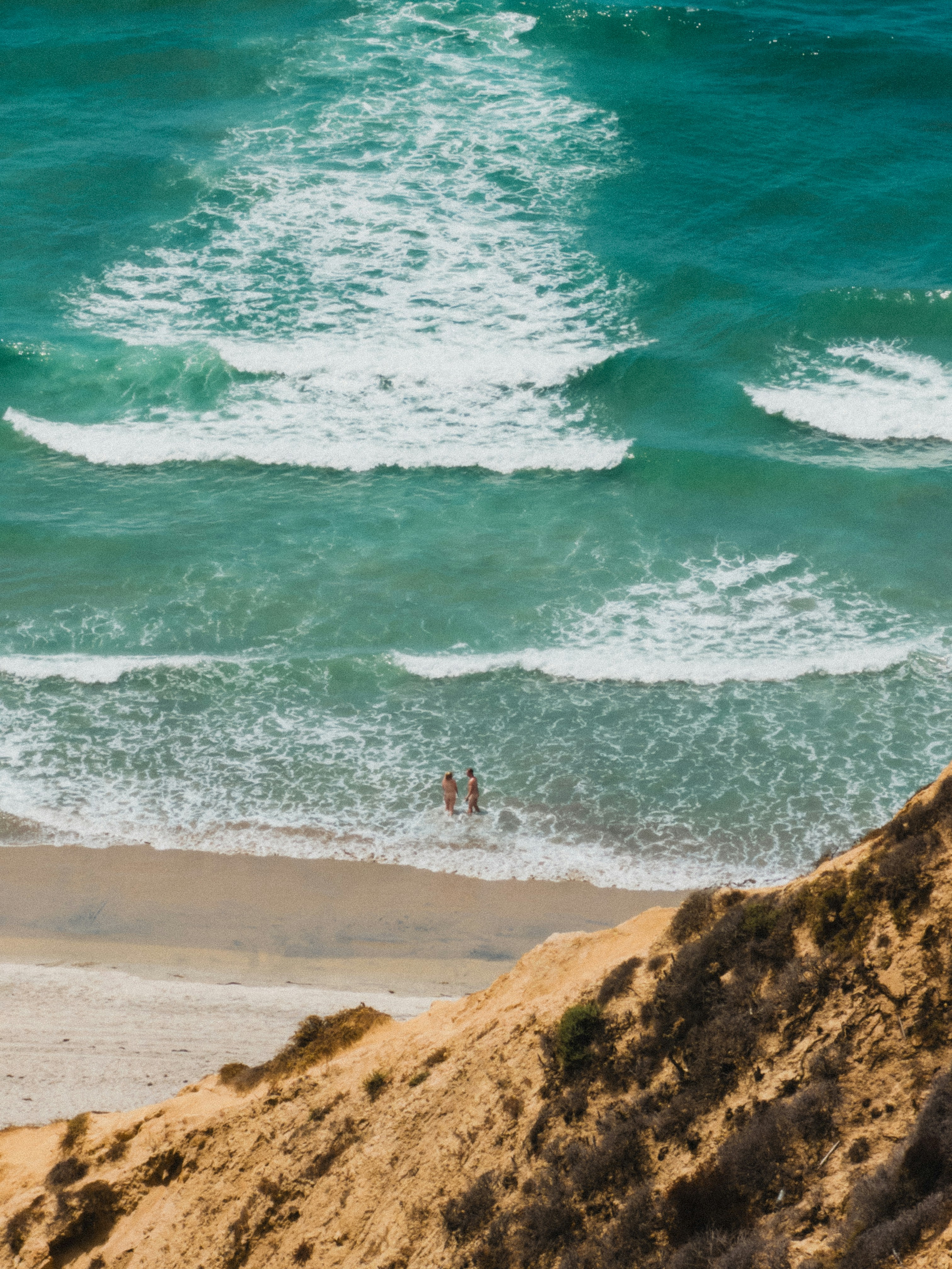 Two people standing in the shallow turquoise surf below a steep sandy cliff