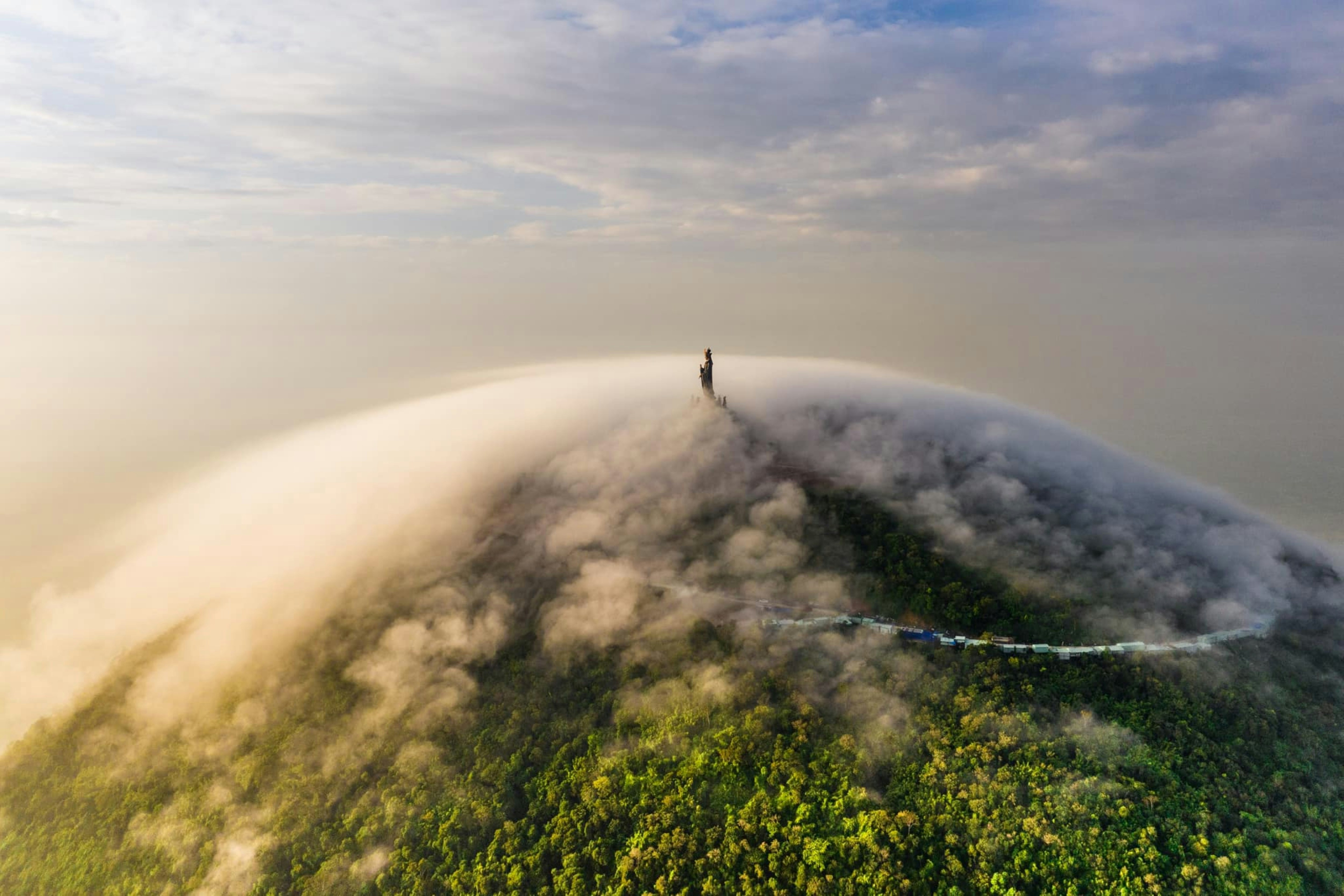 A tall statue on a forested mountain peak surrounded by a thick cloud layer
