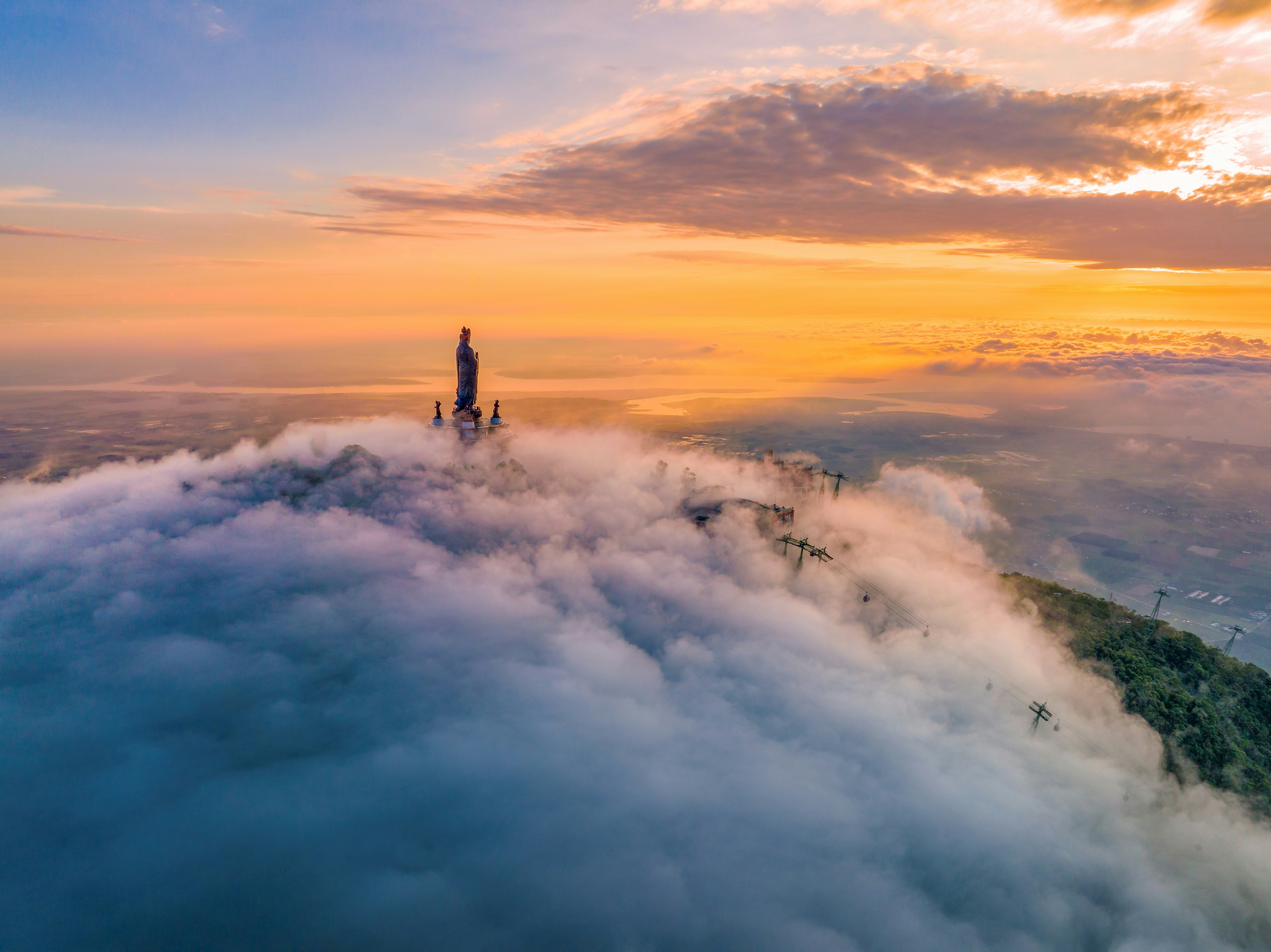 Aerial view of a tall statue on a mountain peak above thick clouds