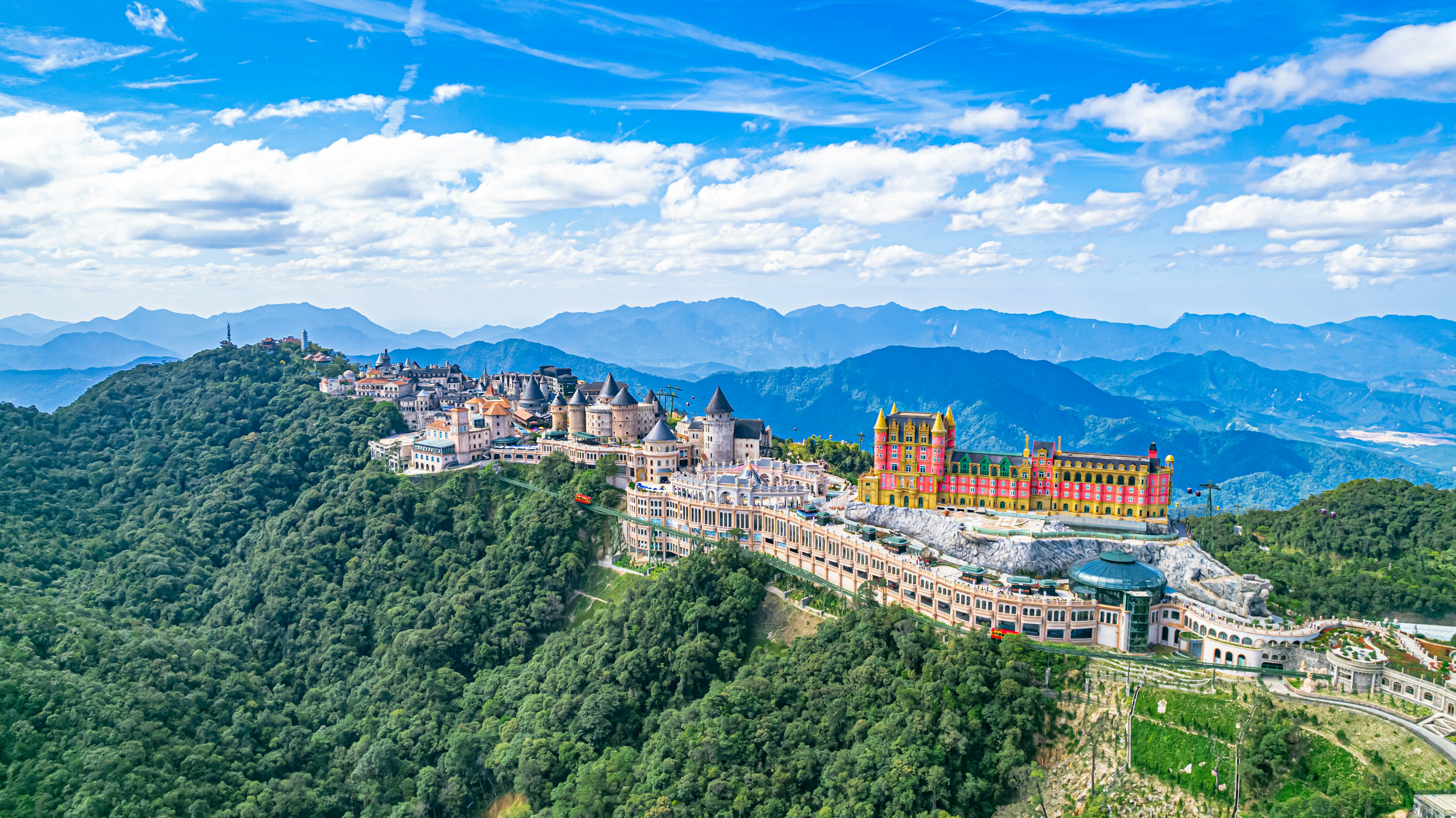 A sprawling castle complex on a forested mountain ridge under a blue sky