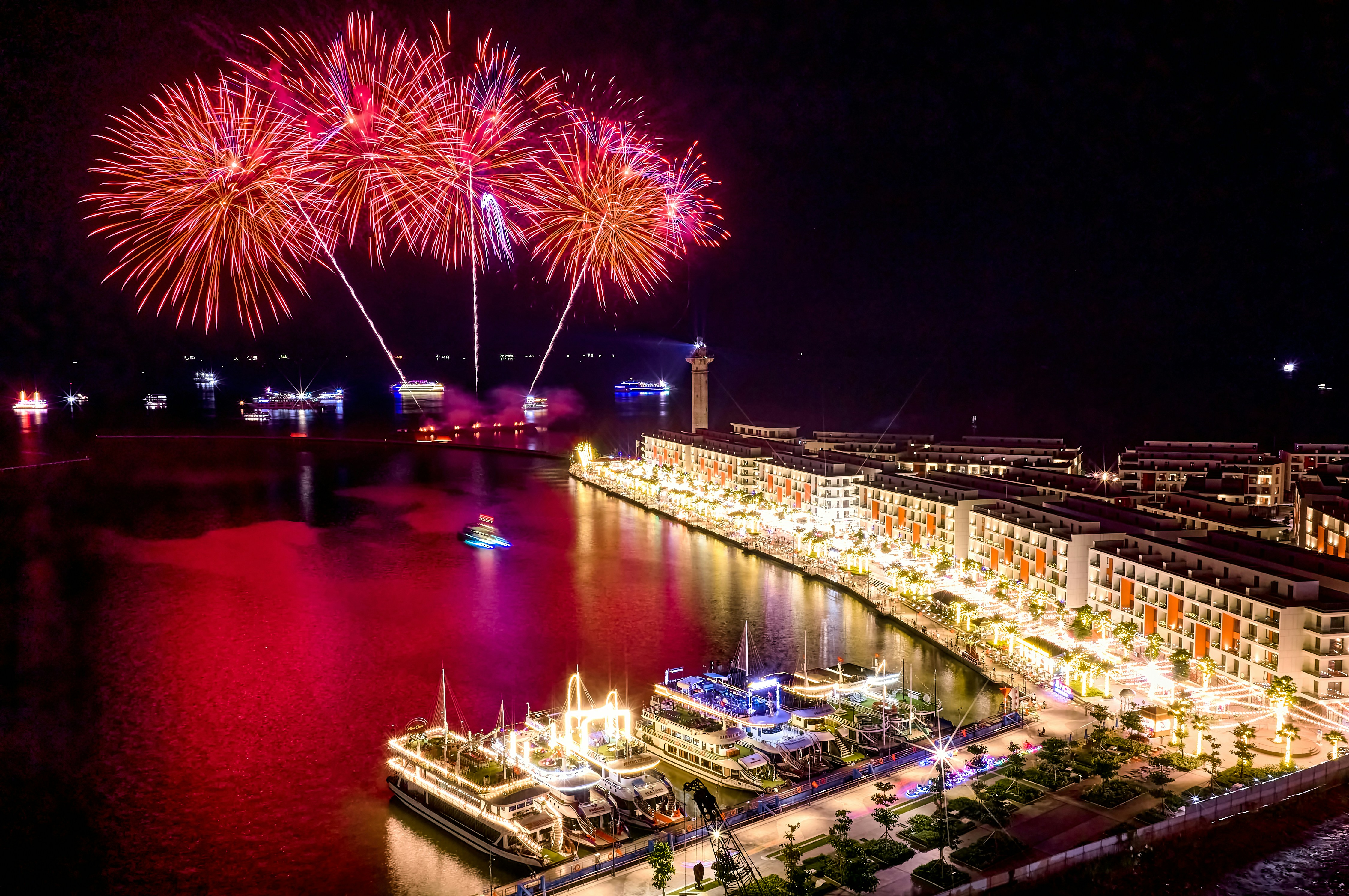 Red fireworks exploding over a harbor with illuminated boats and a coastal resort