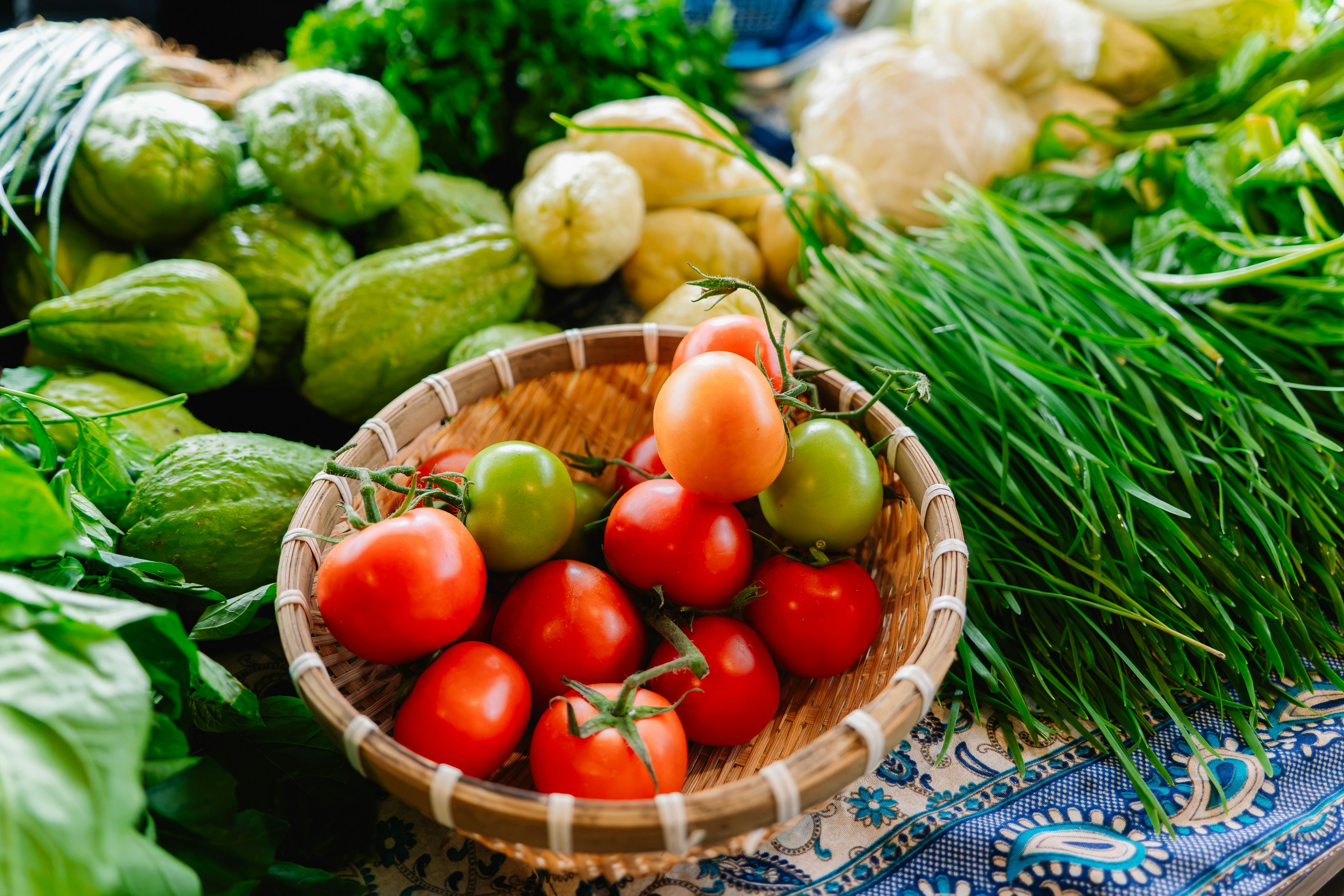 Red and green tomatoes in a woven basket surrounded by fresh vegetables