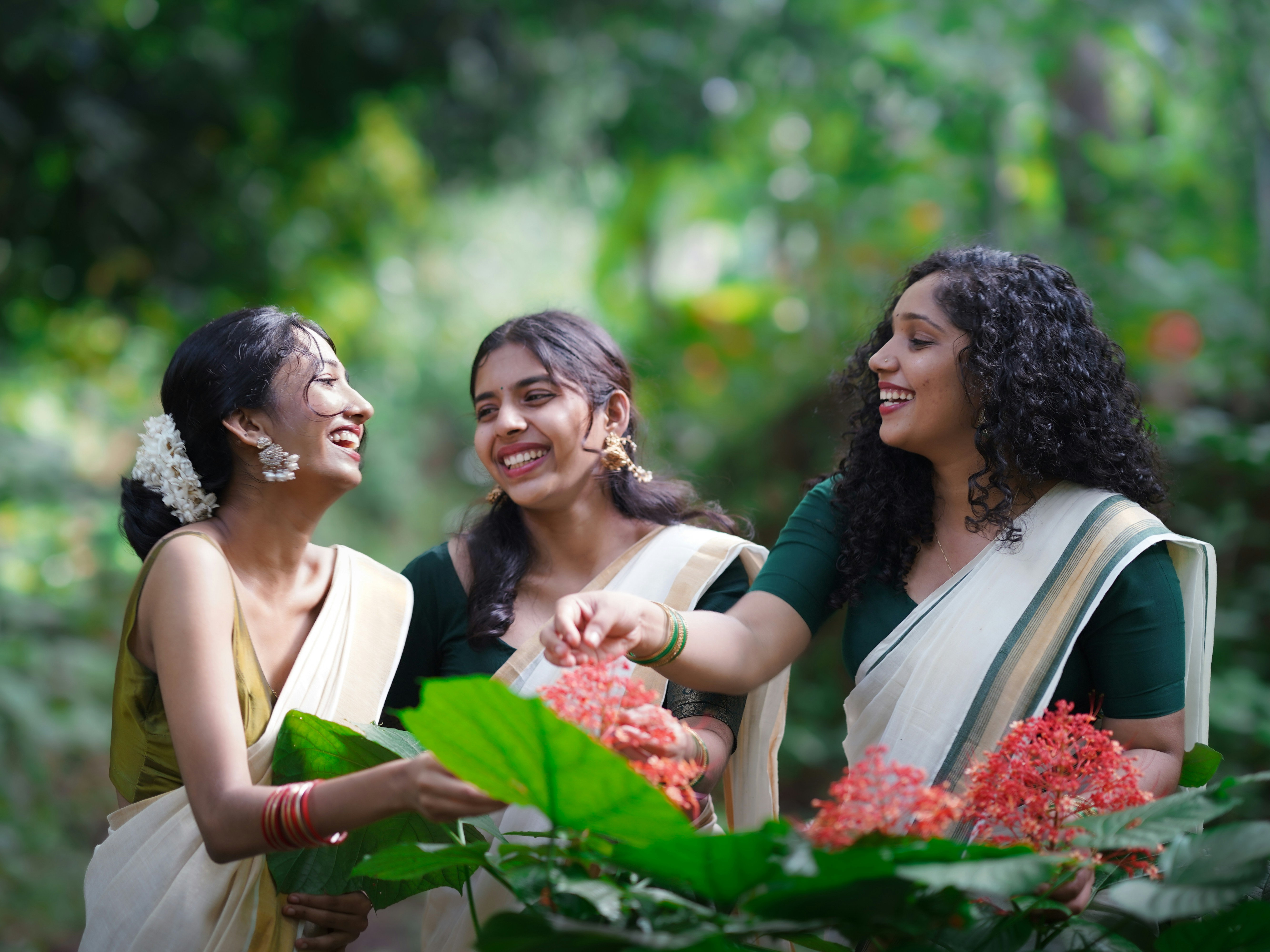 Trois femmes en saris traditionnels du Kerala riant en touchant des fleurs rouges dans un jardin
