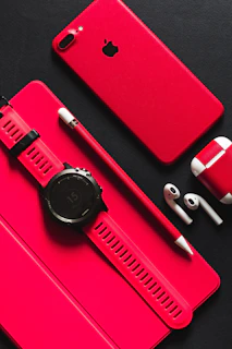 A red smartphone, tablet, stylus, smartwatch, and wireless earbuds on a black surface