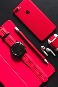 A red smartphone, tablet, stylus, smartwatch, and wireless earbuds on a black surface