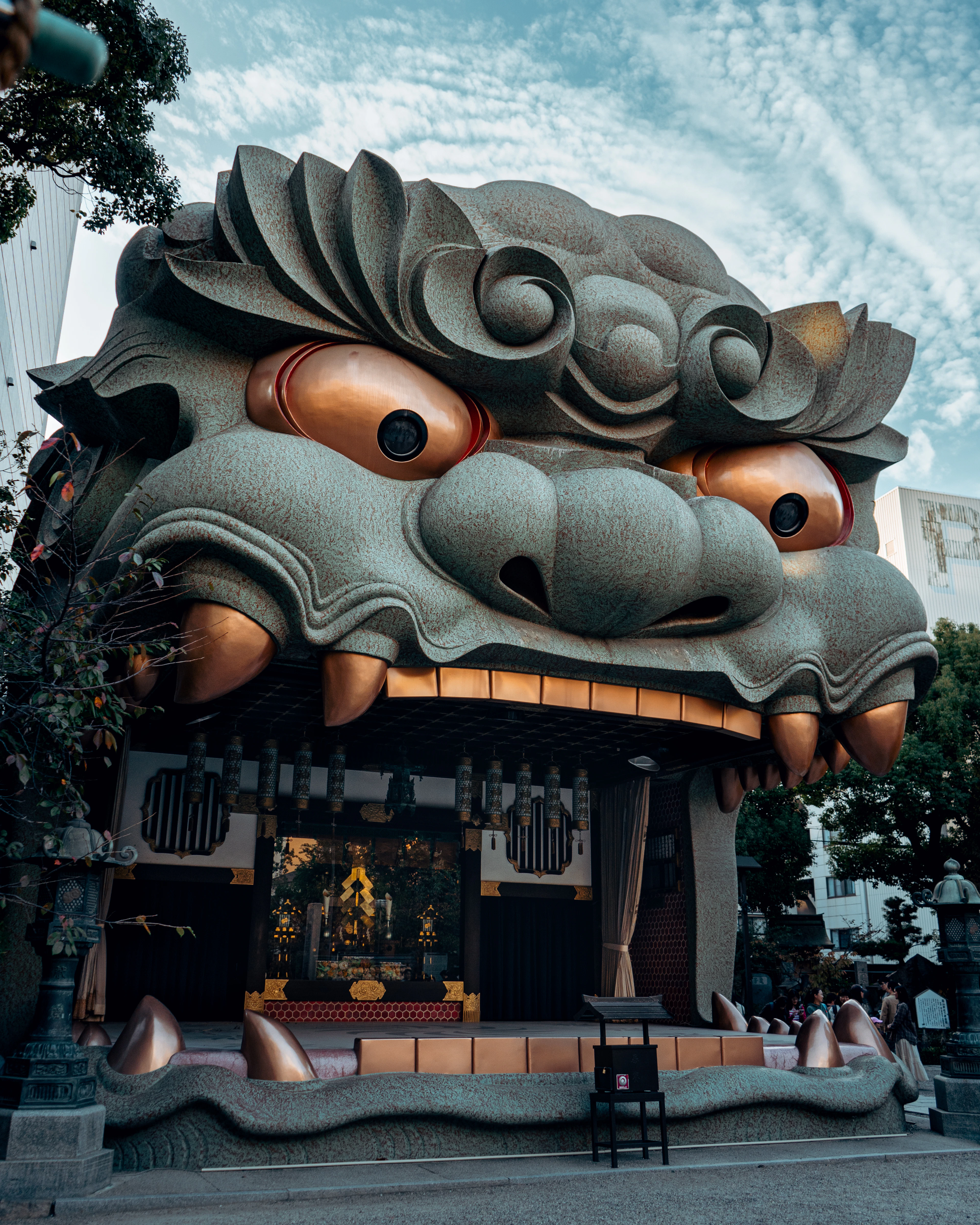 The giant lion-head stage of Namba Yasaka Shrine in Osaka photographed from below, its bronze eyes and teeth catching the light under a sky streaked with high cloud.