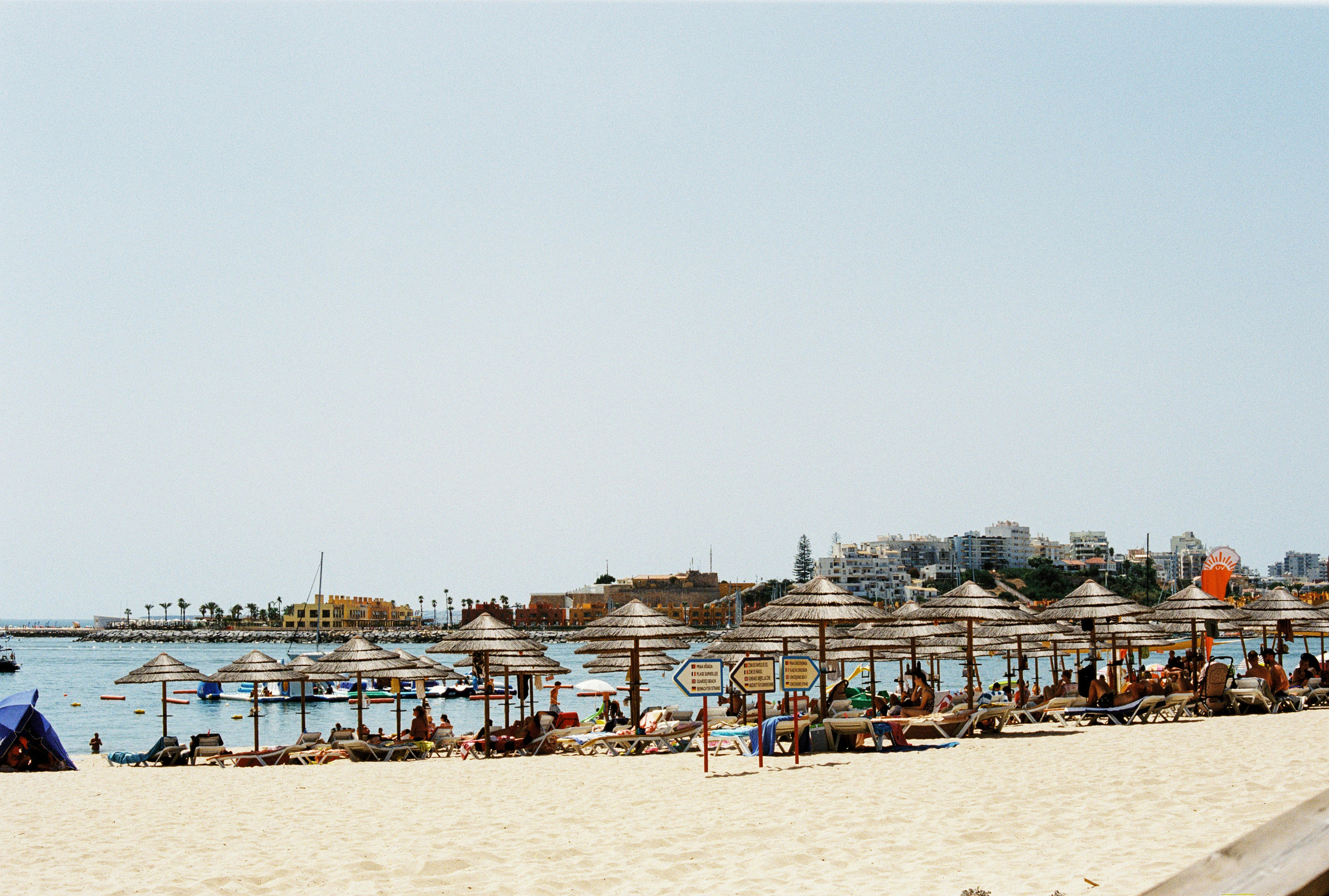 Straw umbrellas and beachgoers on a sandy beach with a coastal town beyond