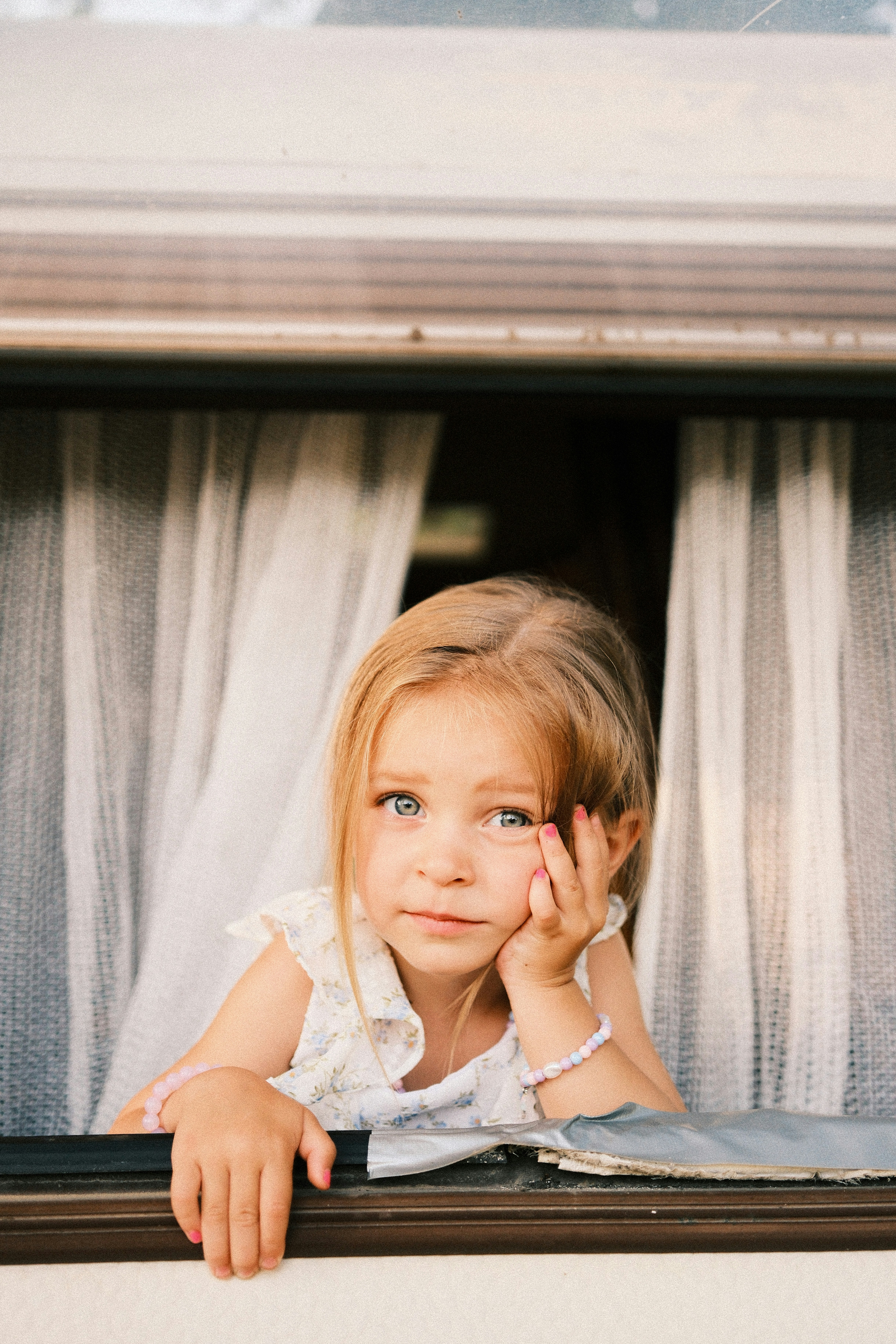 A young child with blonde hair leaning on a window sill with white curtains