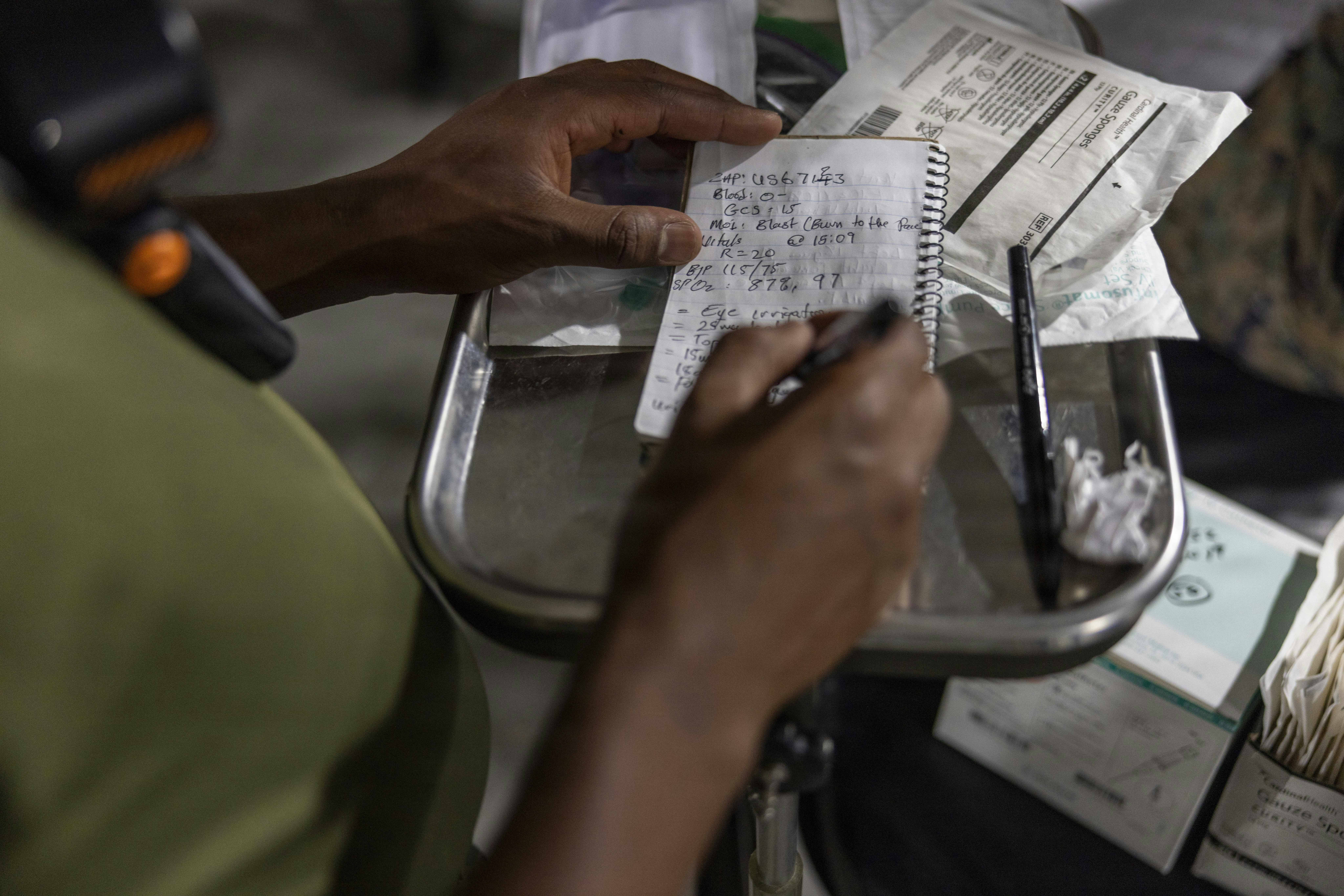A person writing medical notes in a small spiral notebook on a metal tray
