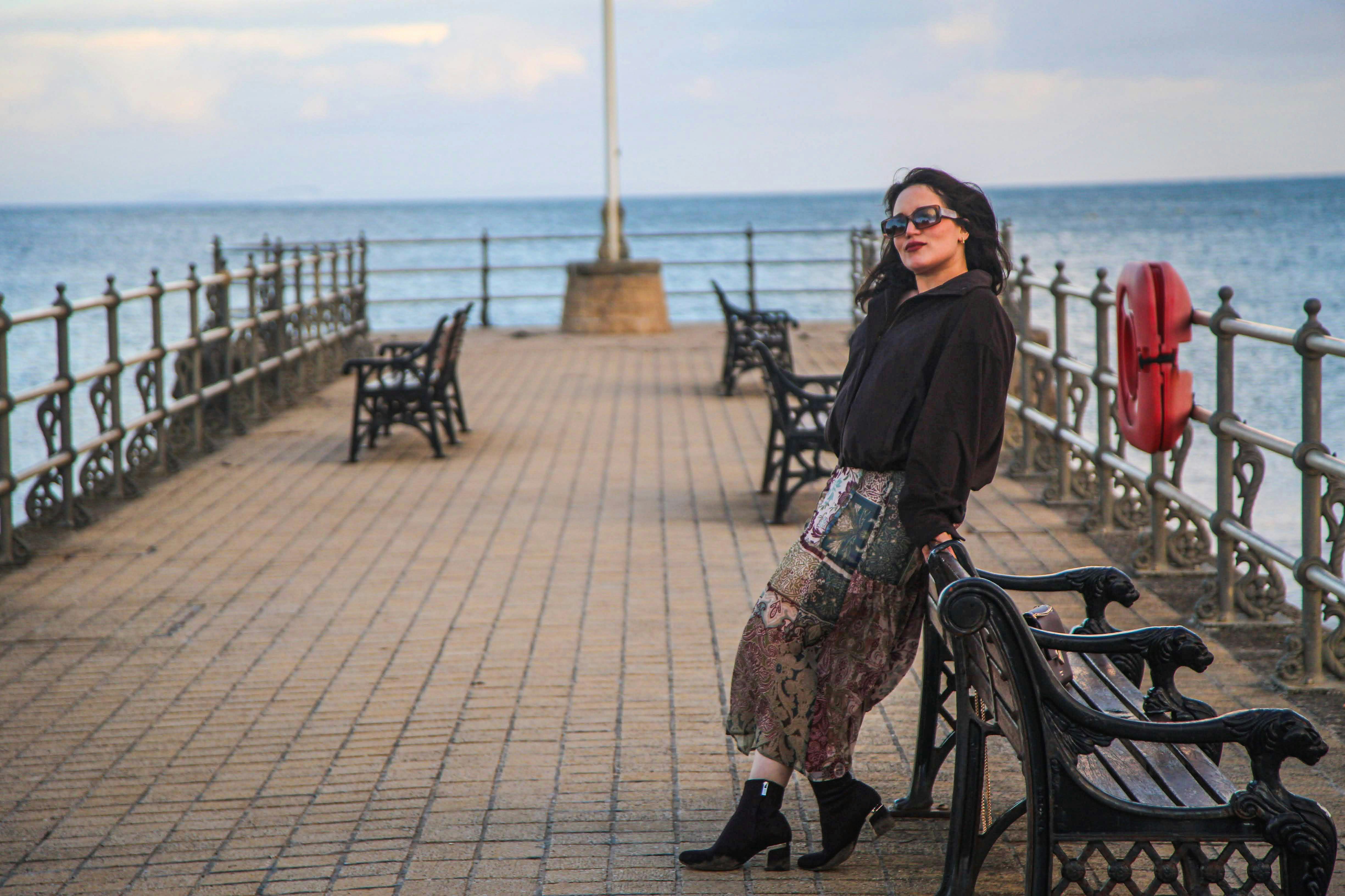 A woman in a black shirt and patterned skirt leaning on a pier bench