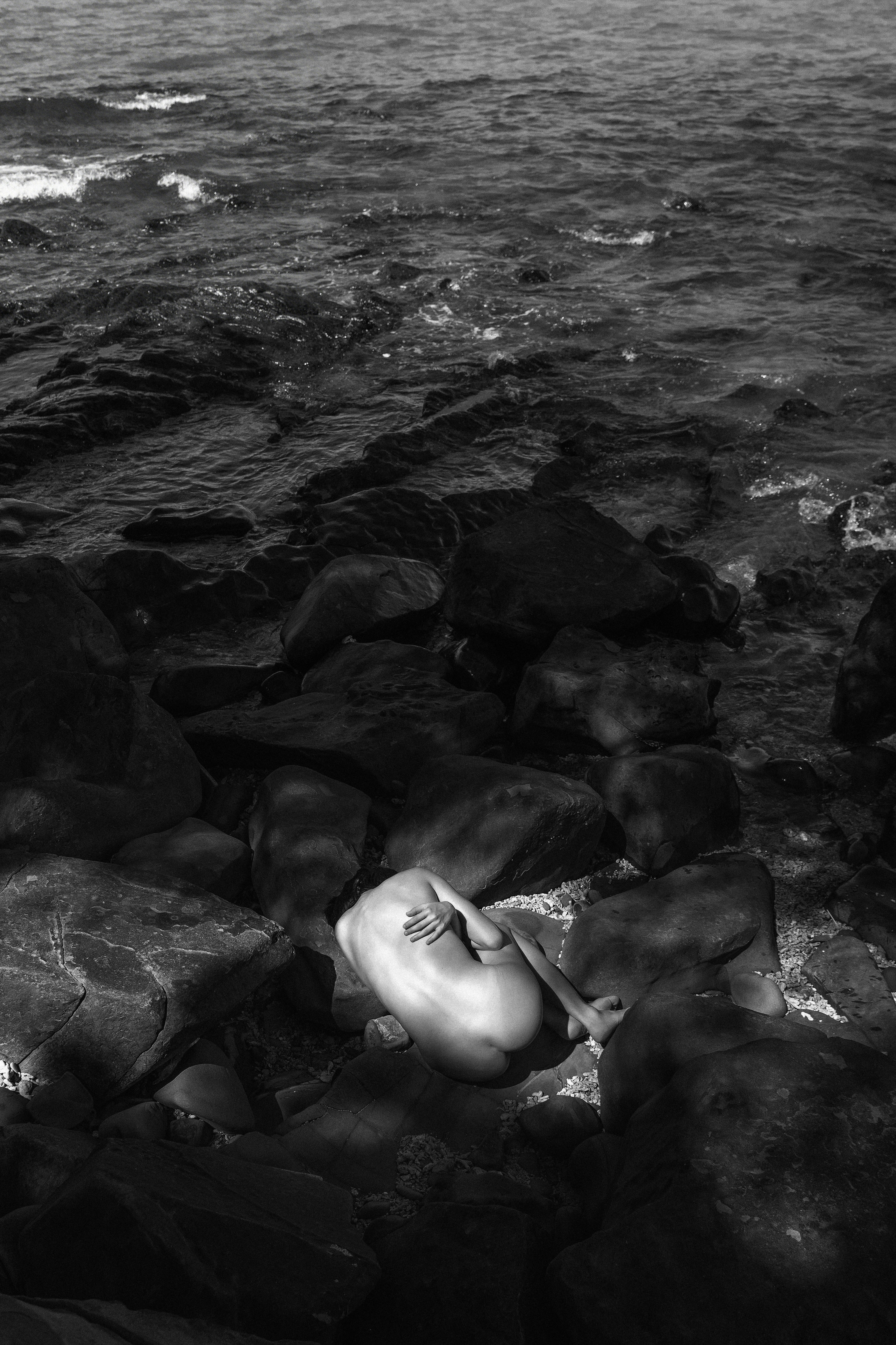 A person curled in a fetal position on dark rocks by the ocean