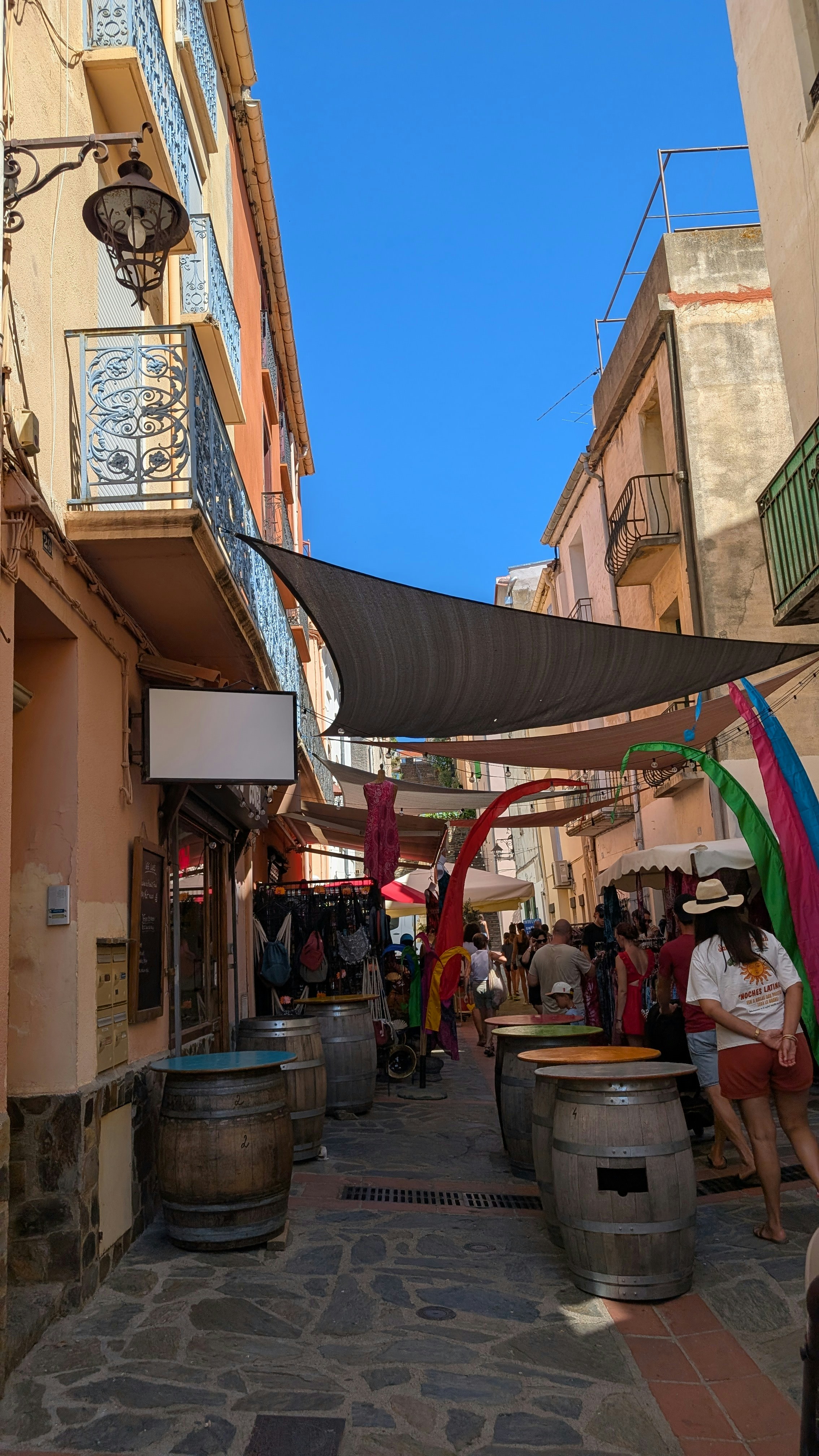 People walking through a narrow street with wooden barrels and overhead shade sails